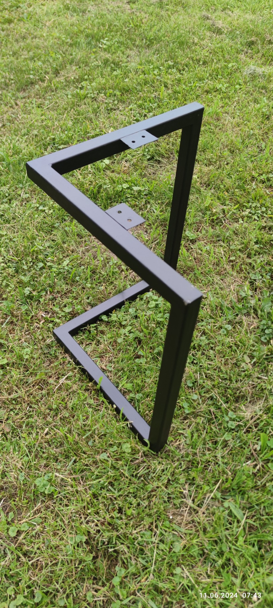 Surowa, spawana konstrukcja metalowa w kształcie litery Z, prawdopodobnie podstawa stołu, leżąca na trawie.