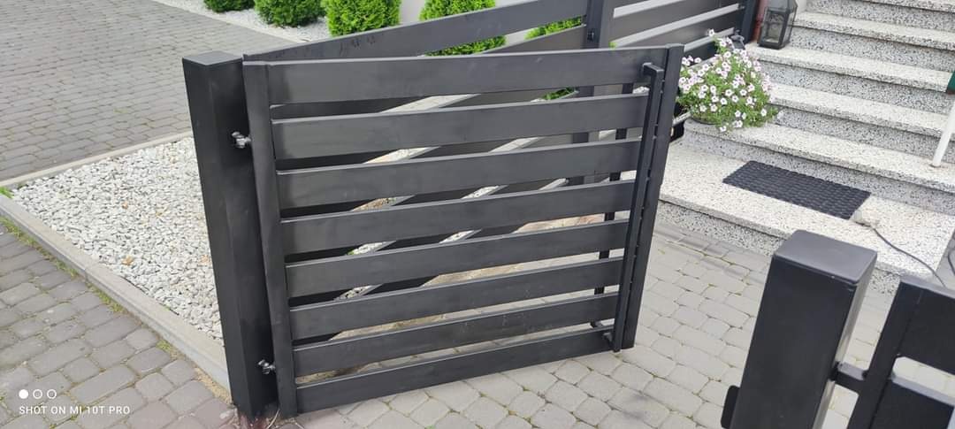 Ciemnoszara furtka ogrodzeniowa z poziomych paneli, zamontowana na posesji z kostki brukowej, widoczne schody wejściowe do domu i dekoracyjne krzewy.