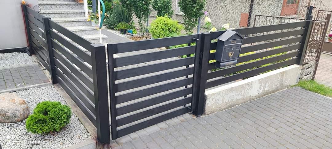 Nowoczesne, poziome, ciemnoszare ogrodzenie z furtką i skrzynką na listy, wykończone betonową podmurówką i kostką brukową, otoczone zielenią.
