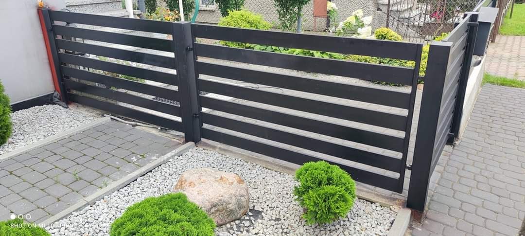 Nowoczesne ogrodzenie z poziomych, czarnych desek metalowych, otoczone białym żwirem, z widocznym fragmentem chodnika z kostki brukowej i dekoracyjnymi krzewami.