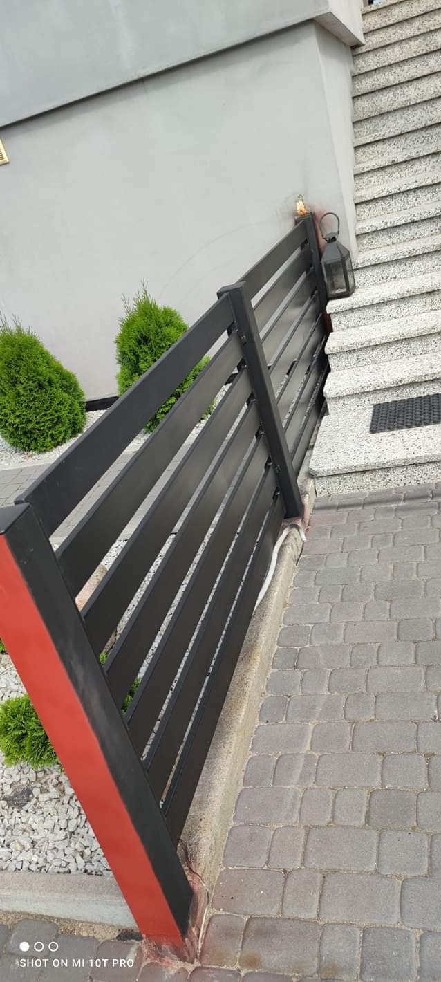 Nowoczesne, czarne ogrodzenie panelowe z czerwonym akcentem na słupku, przylegające do schodów wejściowych z lampą i kostki brukowej.