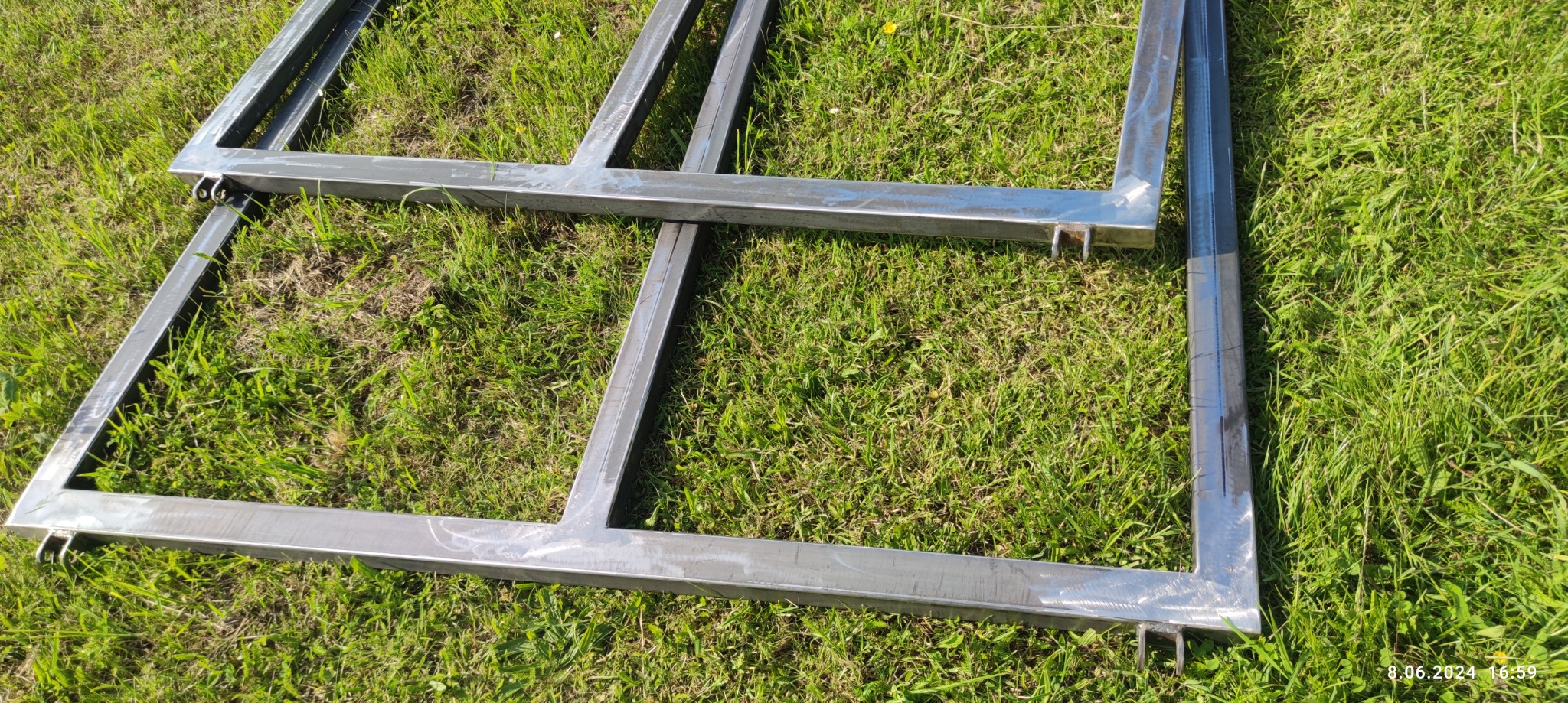 Dwie surowe, spawane metalowe ramy na trawie, prawdopodobnie elementy do ogrodzenia lub bramy, widoczne ślady obróbki metalu.