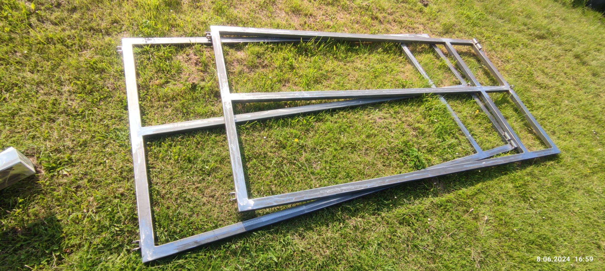 Surowe, spawane metalowe ramy na trawie, prawdopodobnie elementy bramy lub ogrodzenia, z widocznymi zawiasami i śladami po spawaniu.
