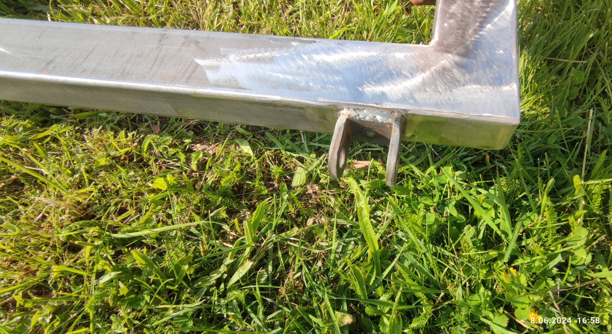 Fragment metalowej ramy z uchwytem, spawana, leżąca na trawie, widoczne ślady obróbki metalu.