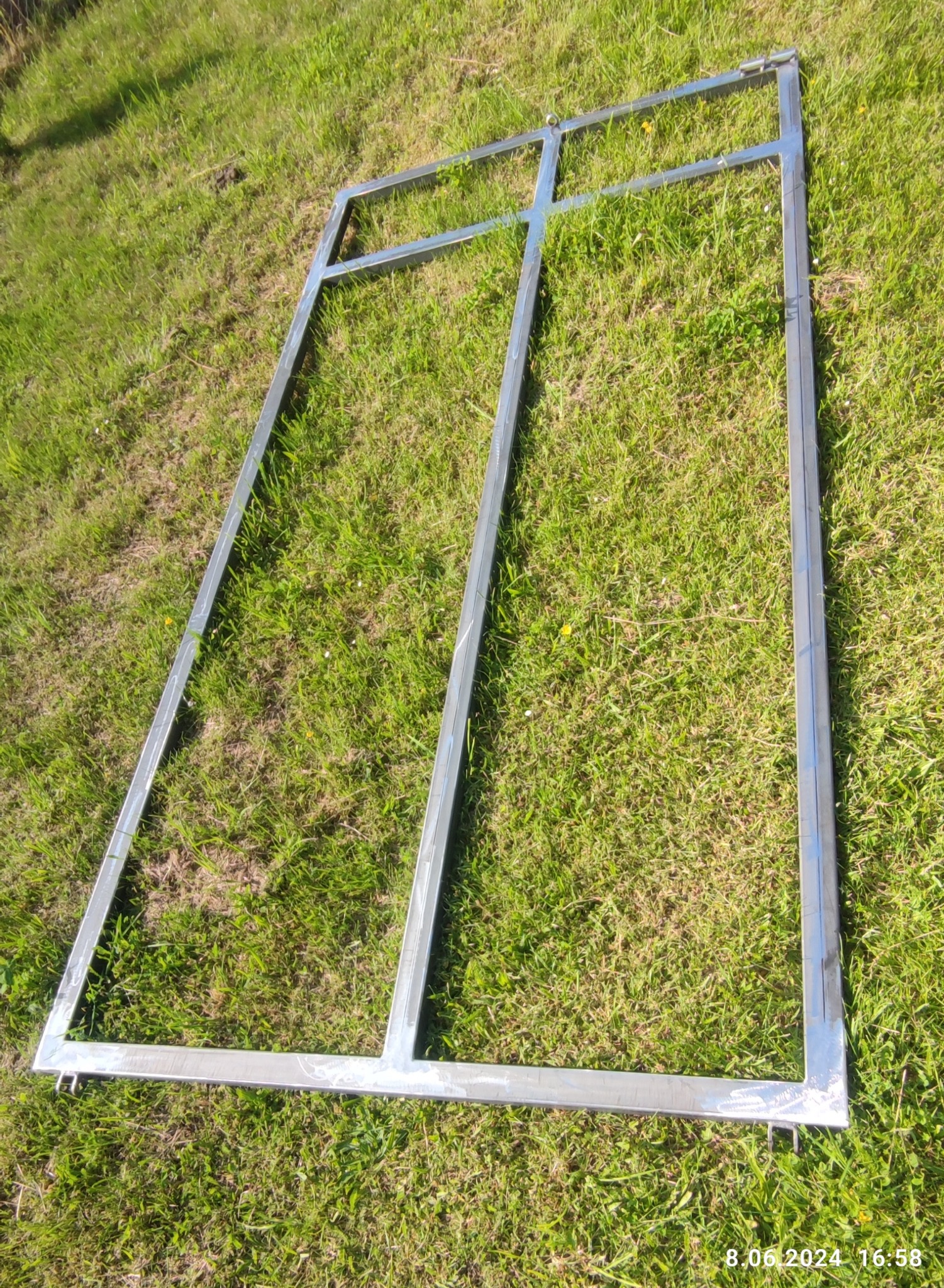 Surowa, spawana, metalowa rama bramy wjazdowej leży na trawie, widoczne spawy i zaczepy, przygotowana do dalszej obróbki.