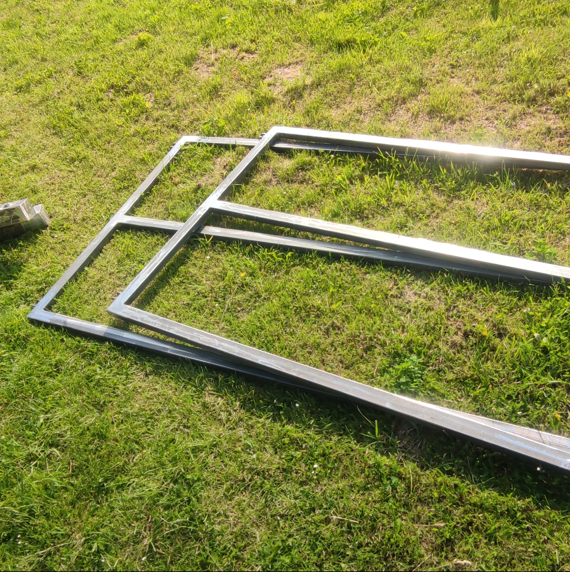 Surowe, metalowe ramy na trawie, prawdopodobnie elementy konstrukcyjne ogrodzenia lub bramy, widoczne spawane łączenia.