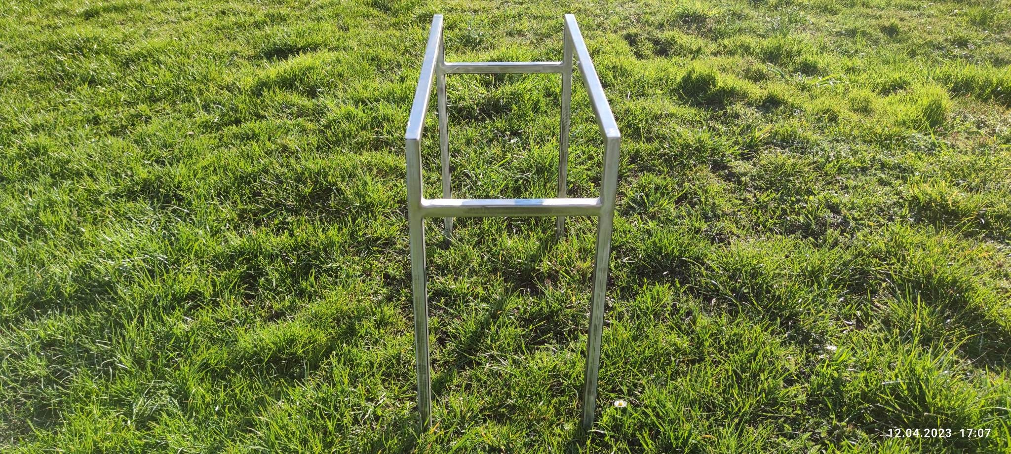 Surowa metalowa konstrukcja, prawdopodobnie szkielet przyszłej pergoli, stojąca na trawniku w słoneczny dzień.