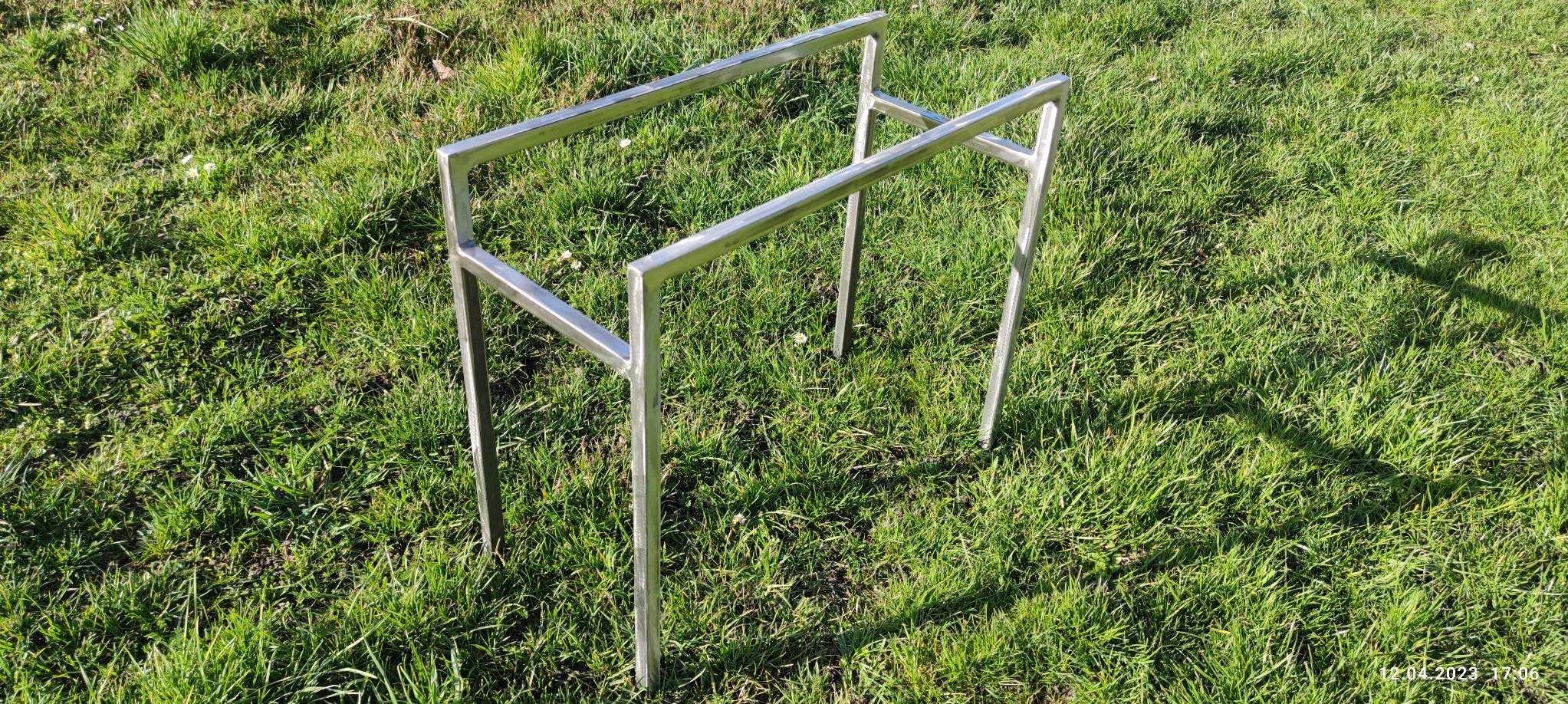 Surowa, metalowa konstrukcja przypominająca stelaż, zespawana z kwadratowych profili, stojąca na trawie w słoneczny dzień.