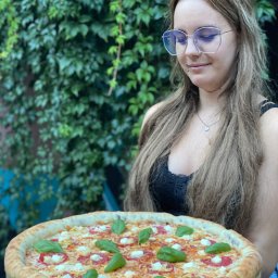 Pizzeria Papay Legionowo - Młoda kobieta z piercingiem w wardze, w okularach z niebieskimi szkłami, trzyma pizzę z mozzarellą, salami i bazylią na drewnianej desce, na tle ściany porośniętej bluszczem.