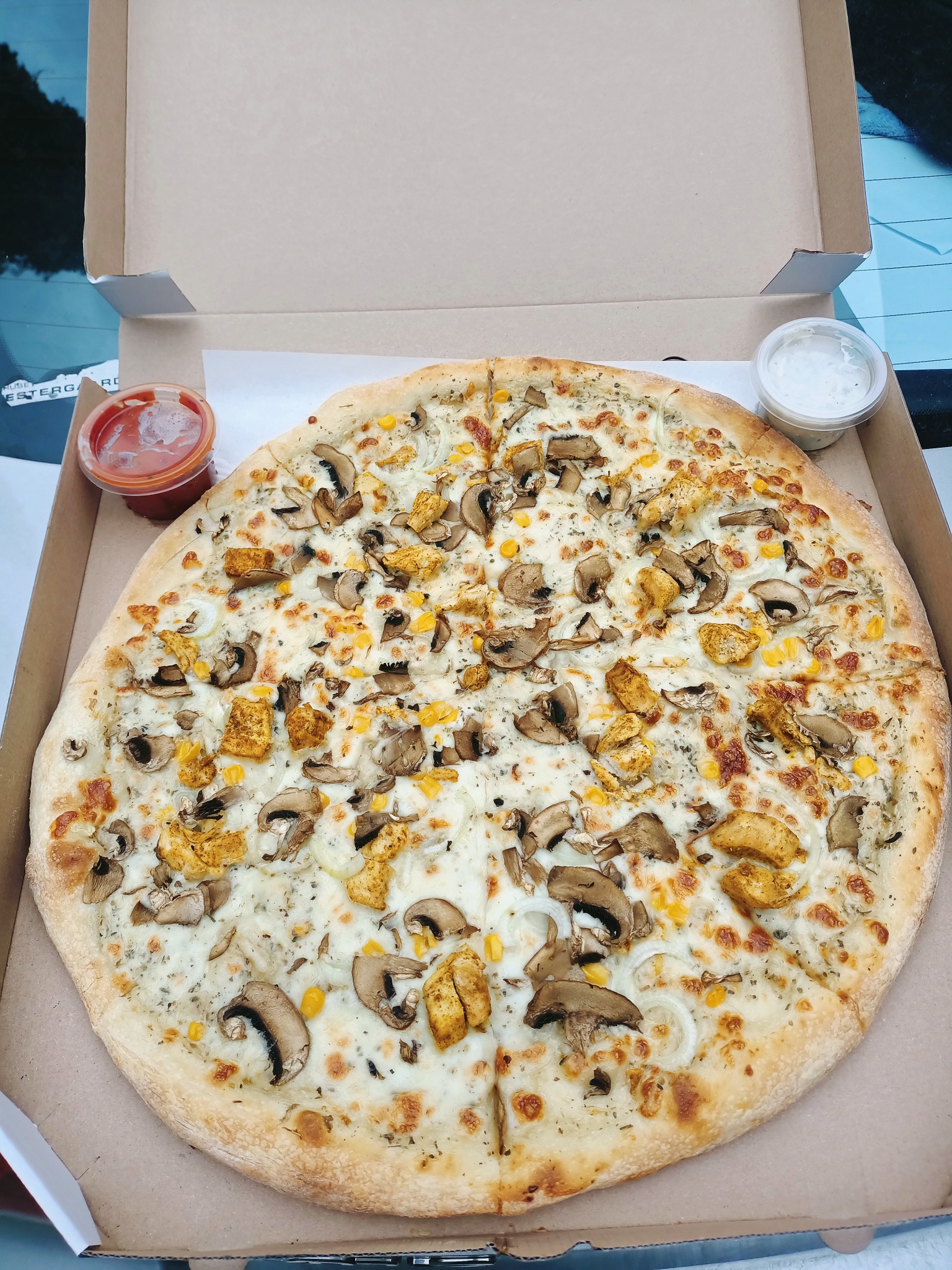 Okrągła pizza z kurczakiem, pieczarkami, kukurydzą i cebulą, pocięta na kawałki, w kartonowym pudełku z dwoma pojemnikami sosów: czerwonym i białym.