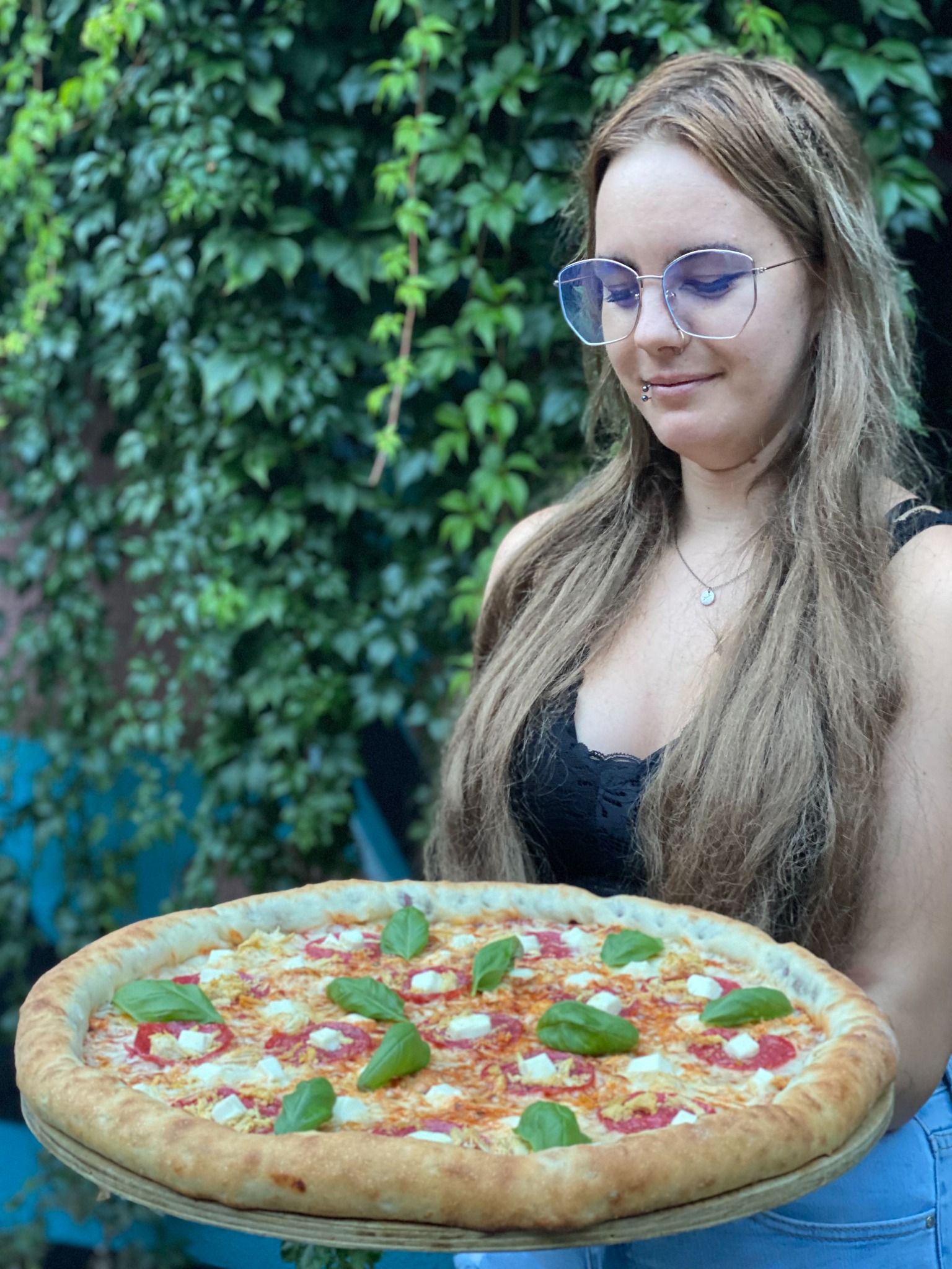 Młoda kobieta z piercingiem w wardze, w okularach z niebieskimi szkłami, trzyma pizzę z mozzarellą, salami i bazylią na drewnianej desce, na tle ściany porośniętej bluszczem.