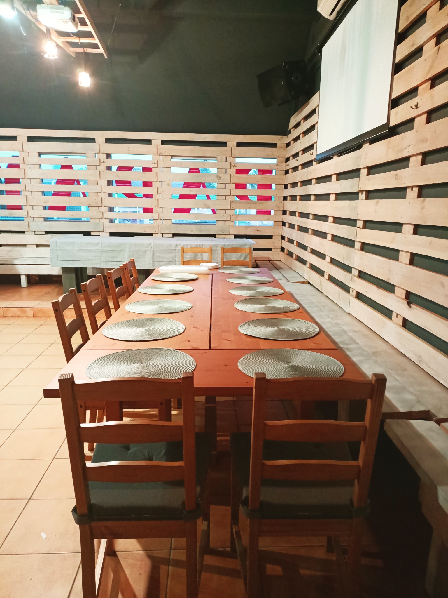 Długi drewniany stół z krzesłami i okrągłymi podkładkami, przygotowany do uroczystości w sali z drewnianymi ścianami i projektorem.