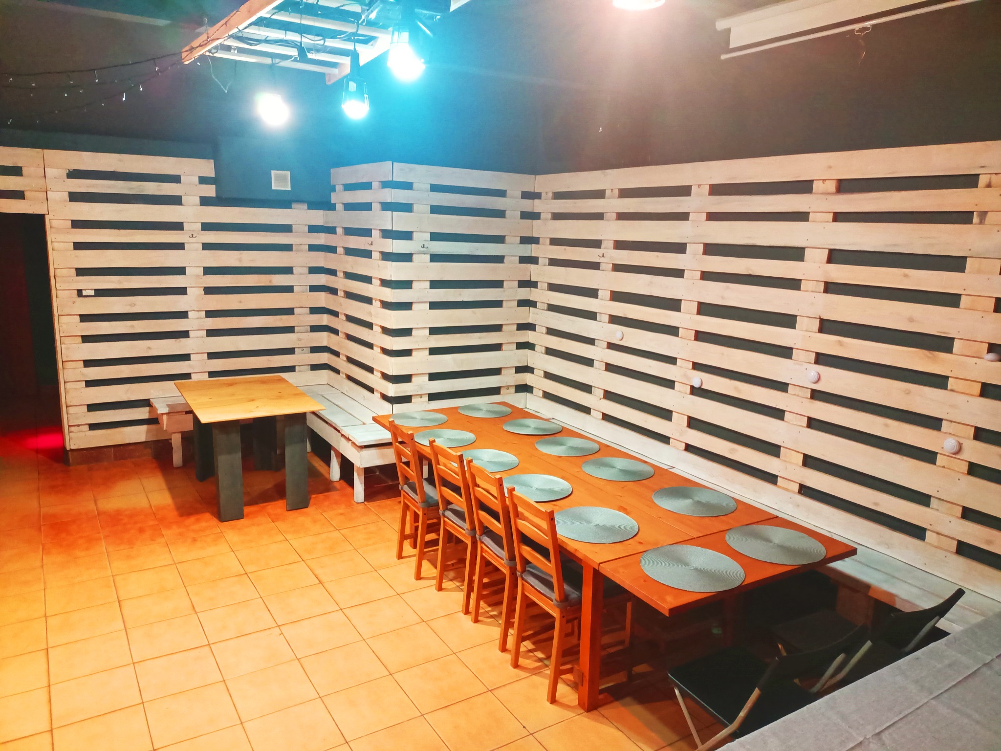 Sala z długim drewnianym stołem nakrytym podkładkami, krzesłami i ścianami wyłożonymi jasnymi paletami, oświetlona przez jasne żarówki.