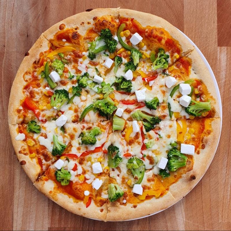 Pizza wegetariańska z brokułami, kukurydzą, papryką i kostkami sera feta, ułożona na drewnianym blacie.