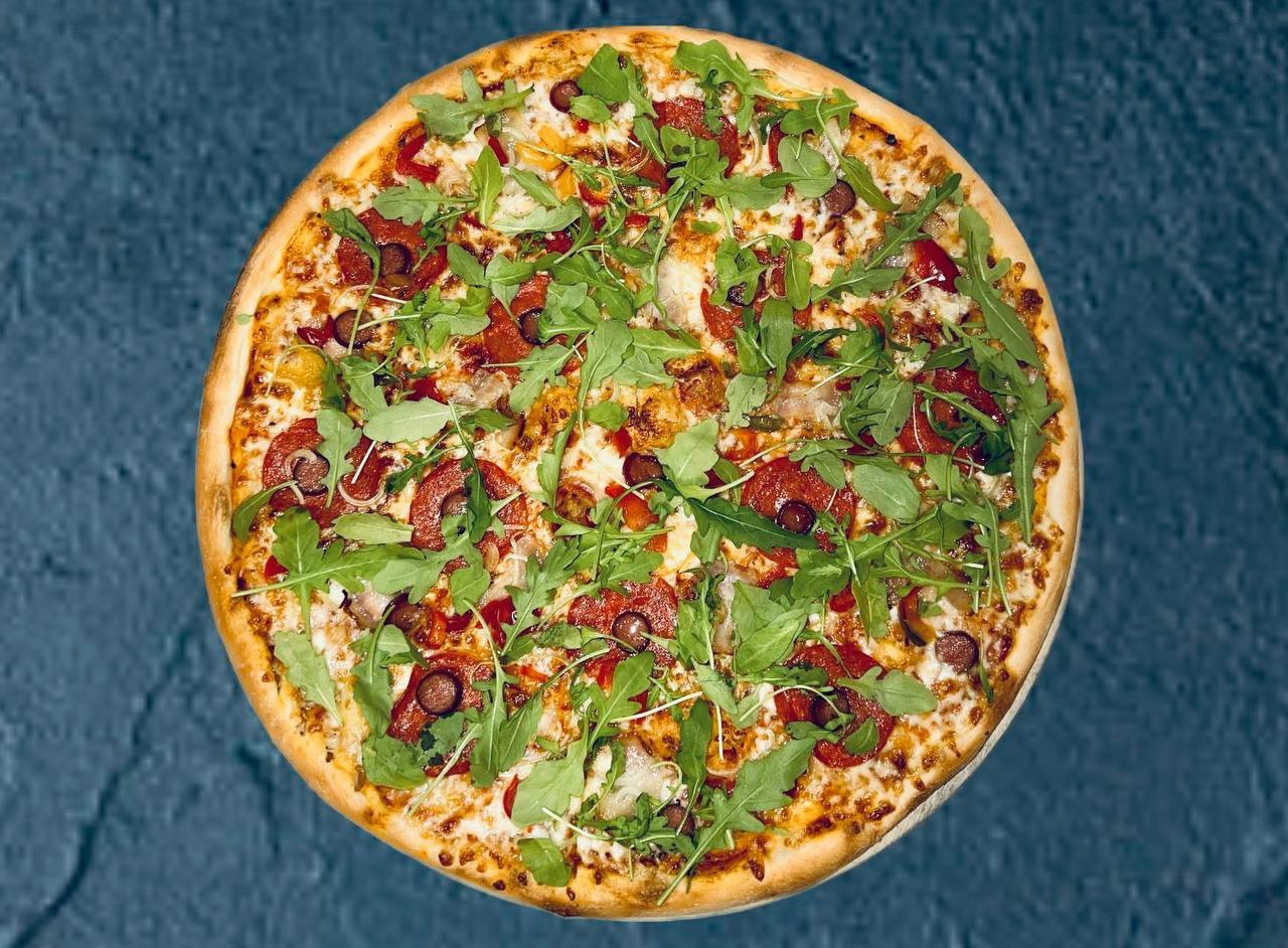 Okrągła pizza z salami, rukolą, papryką i serem na cienkim cieście, ułożona na ciemnym tle.