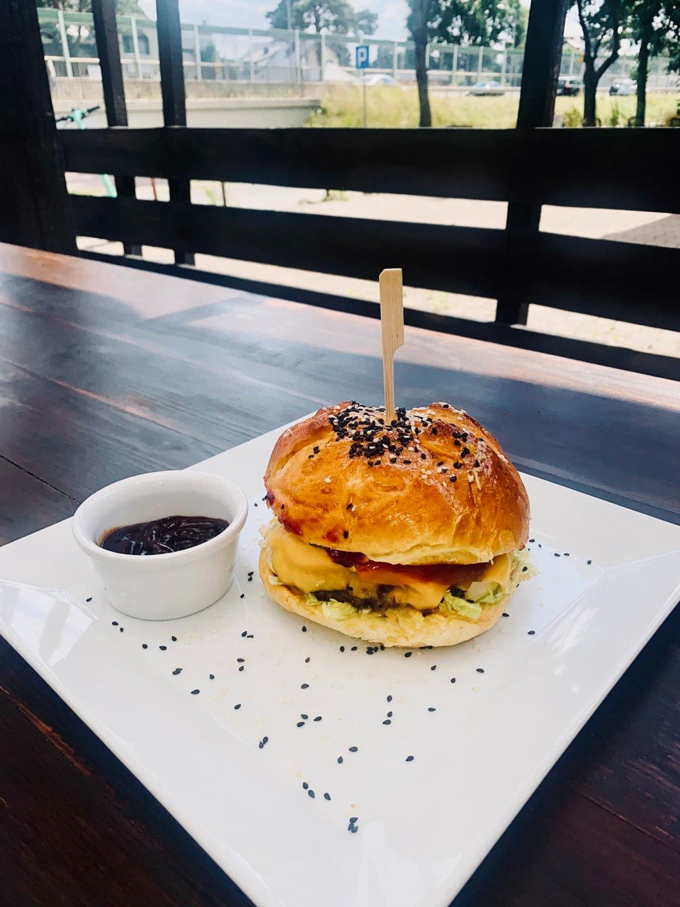 Apetyczny burger z serem i sezamem na białym talerzu obok sosu w białej miseczce, podany na ciemnym drewnianym stole z widokiem na ulicę w tle.