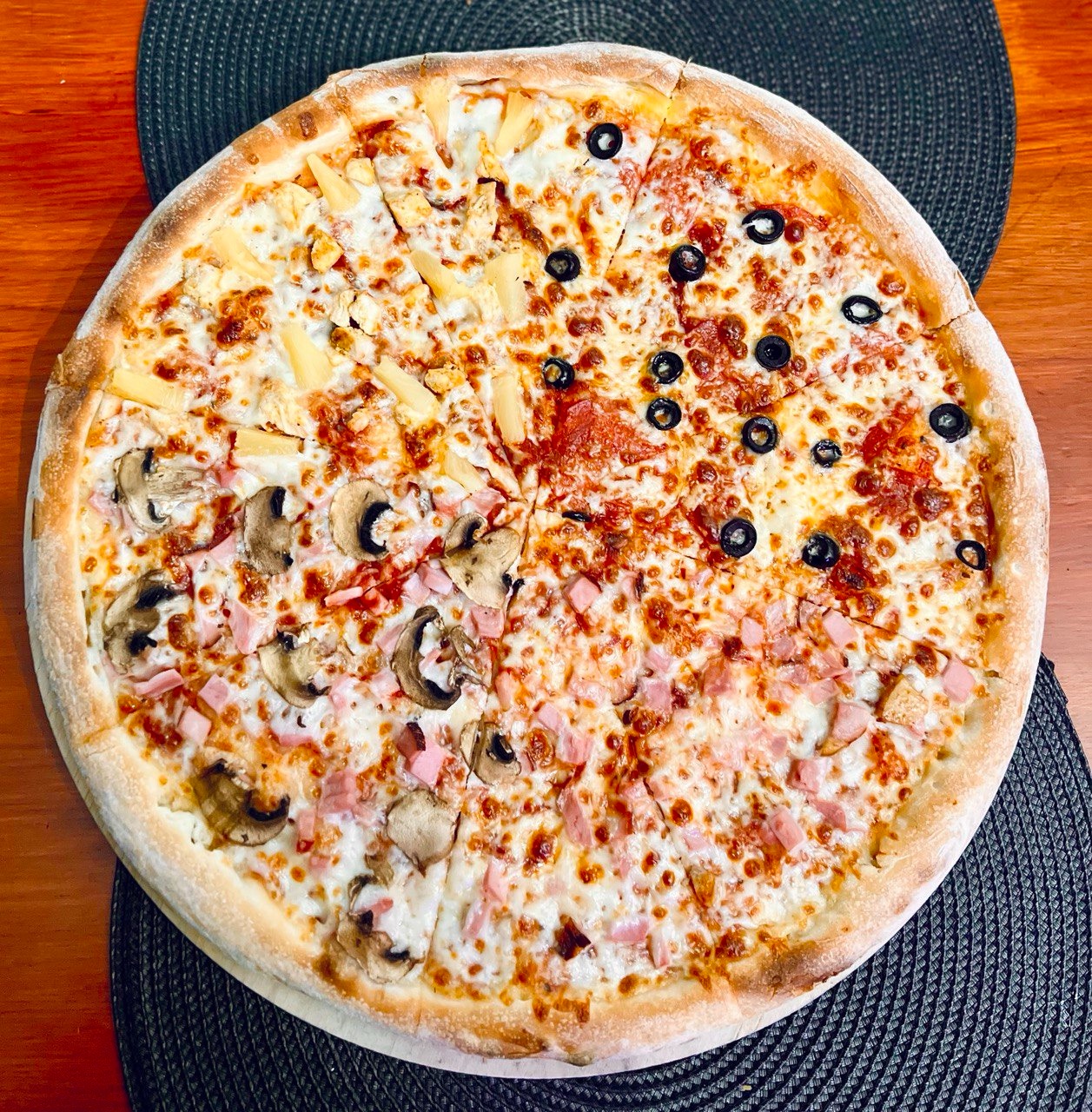 Pizza podzielona na cztery różne smaki: z ananasem i kurczakiem, z salami i czarnymi oliwkami, z pieczarkami i szynką, oraz z szynką. Widok z góry na okrągłym, ciemnym podkładzie.