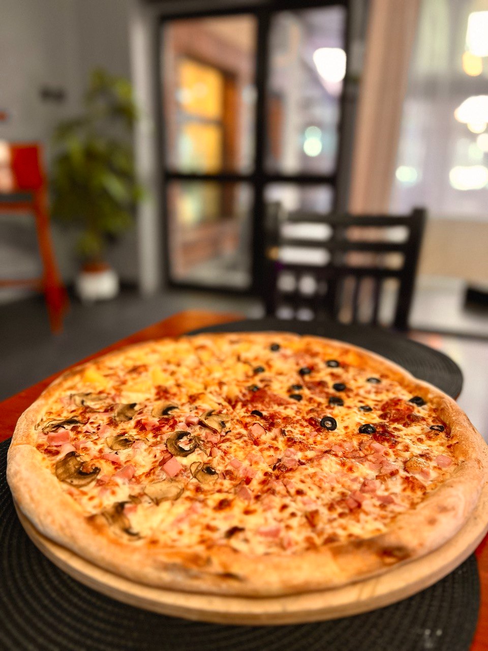 Pizza podzielona na dwie części: jedna z pieczarkami i szynką, druga z oliwkami i salami, na drewnianej desce, w tle rozmyte wnętrze lokalu.