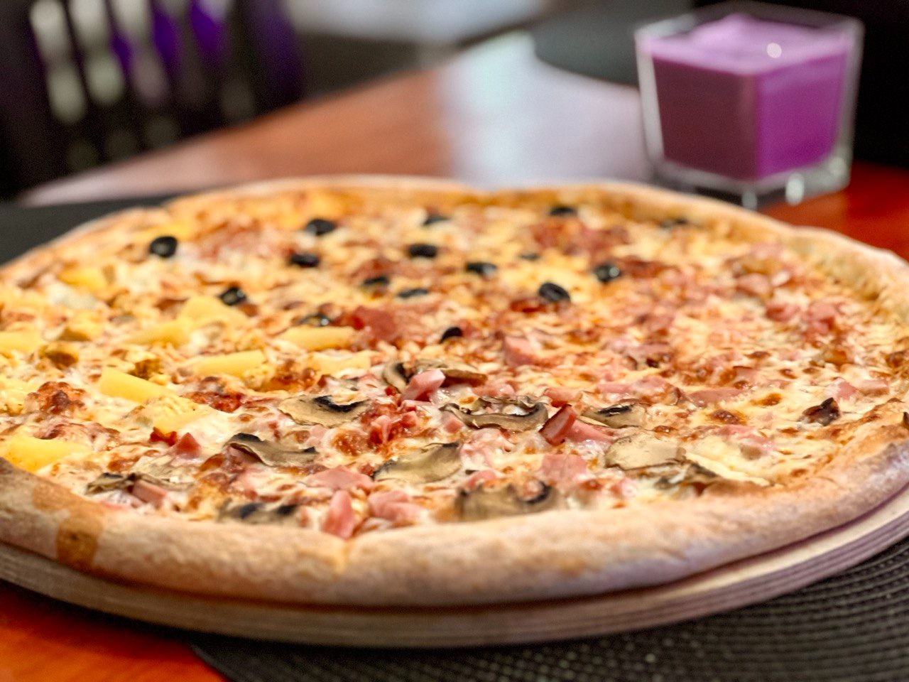 Pizza podzielona na cztery smaki: ananas, oliwki, pieczarki i szynka, podana na drewnianej desce, w tle fioletowy napój w szklance.