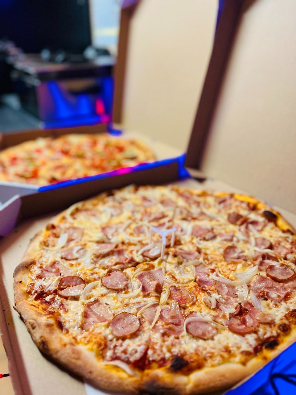 Pizza z kiełbasą i cebulą w kartonie, widoczne plastikowe podpórki, w tle druga pizza w pudełku, ujęcie z bliska.