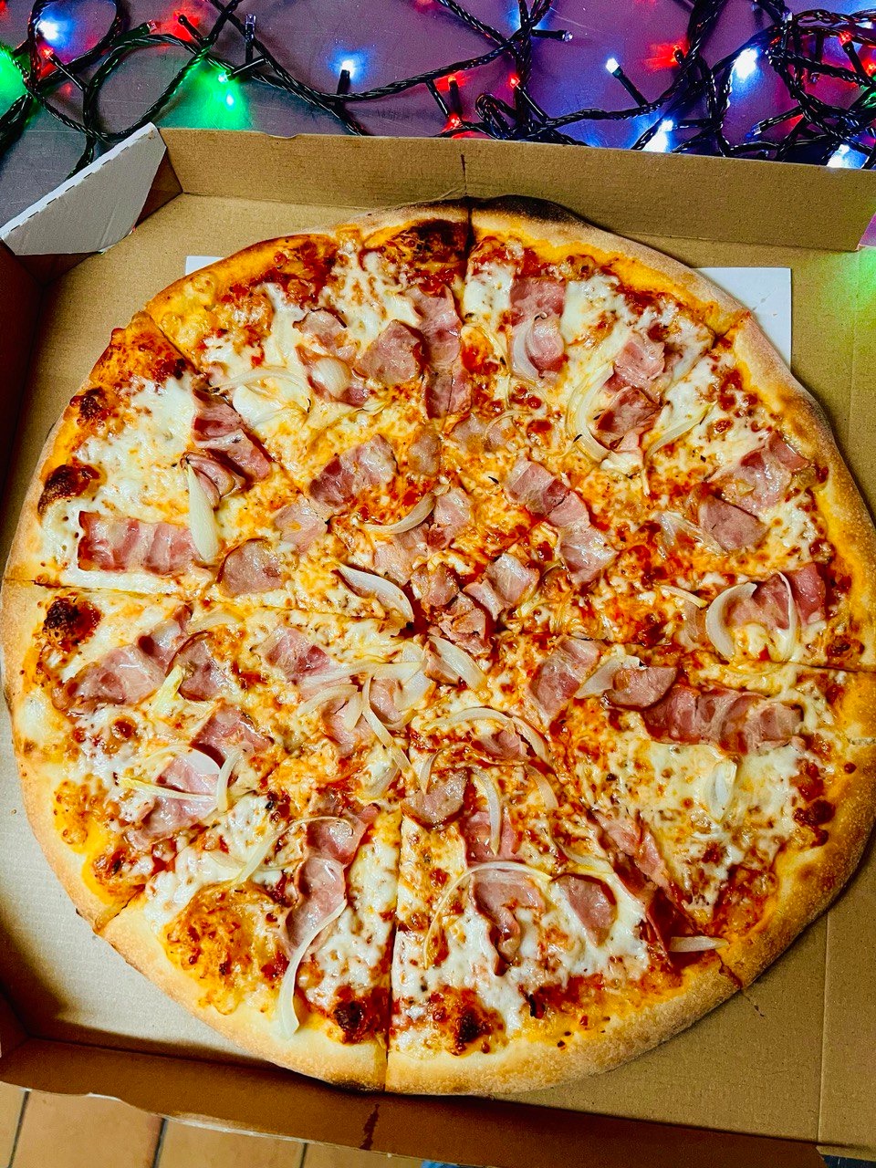 Okrągła pizza z szynką i cebulą, pokrojona na kawałki, w kartonowym pudełku, z rozmytym tłem z lampek choinkowych.