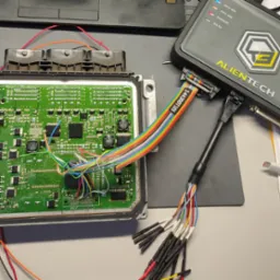 Otwarty moduł sterujący silnika (ECU) z podłączonymi kolorowymi przewodami do programatora Alientech, widoczne diody sygnalizacyjne urządzenia diagnostycznego oraz elementy elektroniczne na płytce...