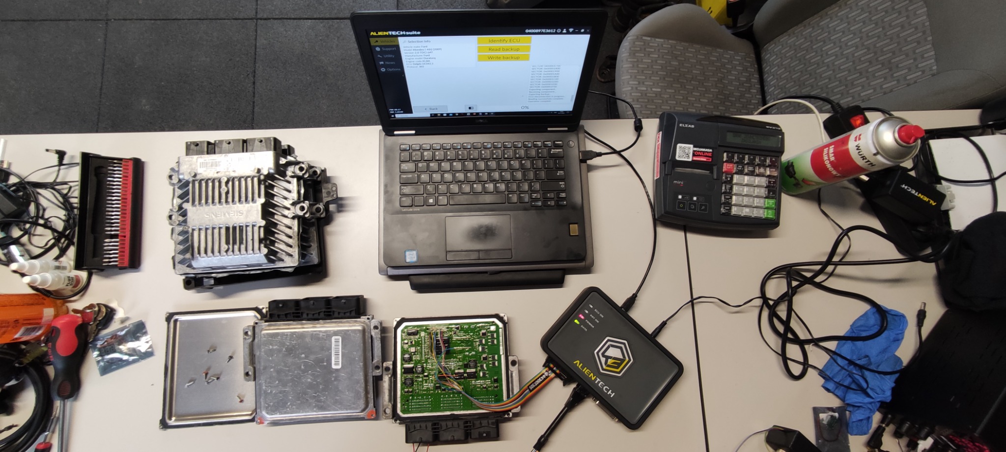 Diagnostyka sterownika silnika samochodowego z użyciem interfejsu diagnostycznego Alientech podłączonego do laptopa z uruchomionym oprogramowaniem, obok widoczne rozłożone elementy sterownika...