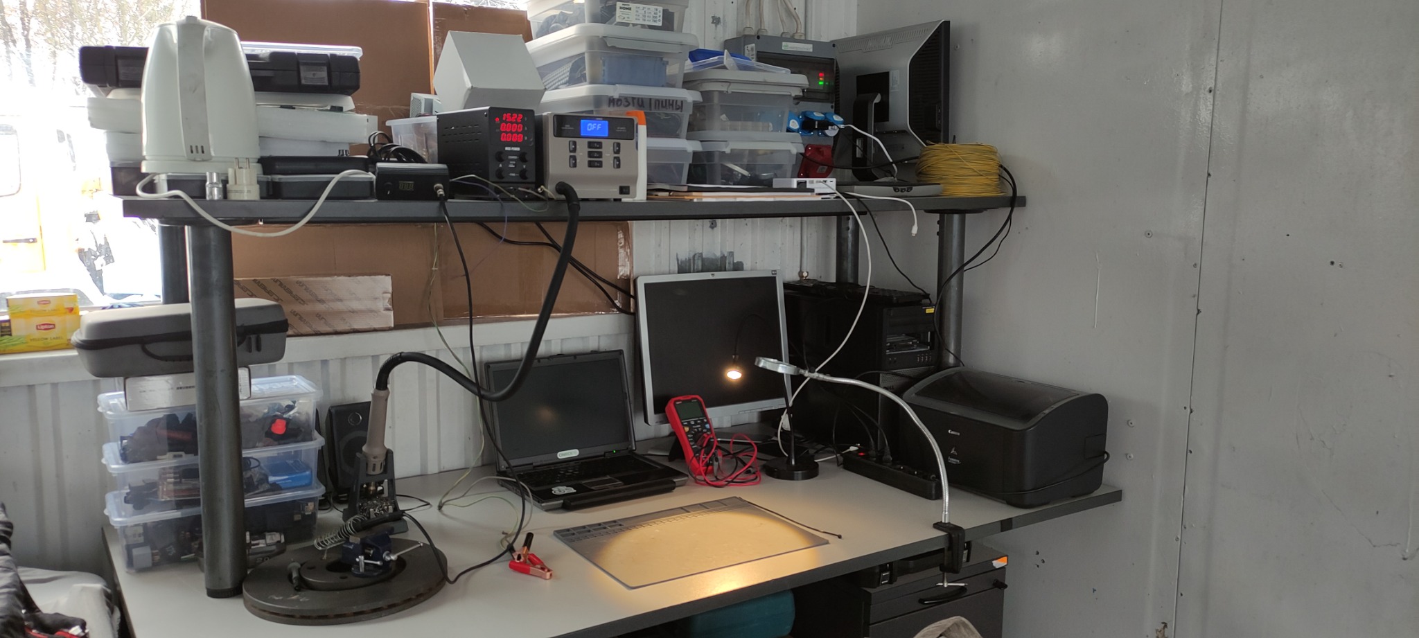 Stanowisko pracy z lutownicą na statywie, komputerem, multimetrem i zasilaczem warsztatowym, umieszczone na metalowym regale w kontenerze.