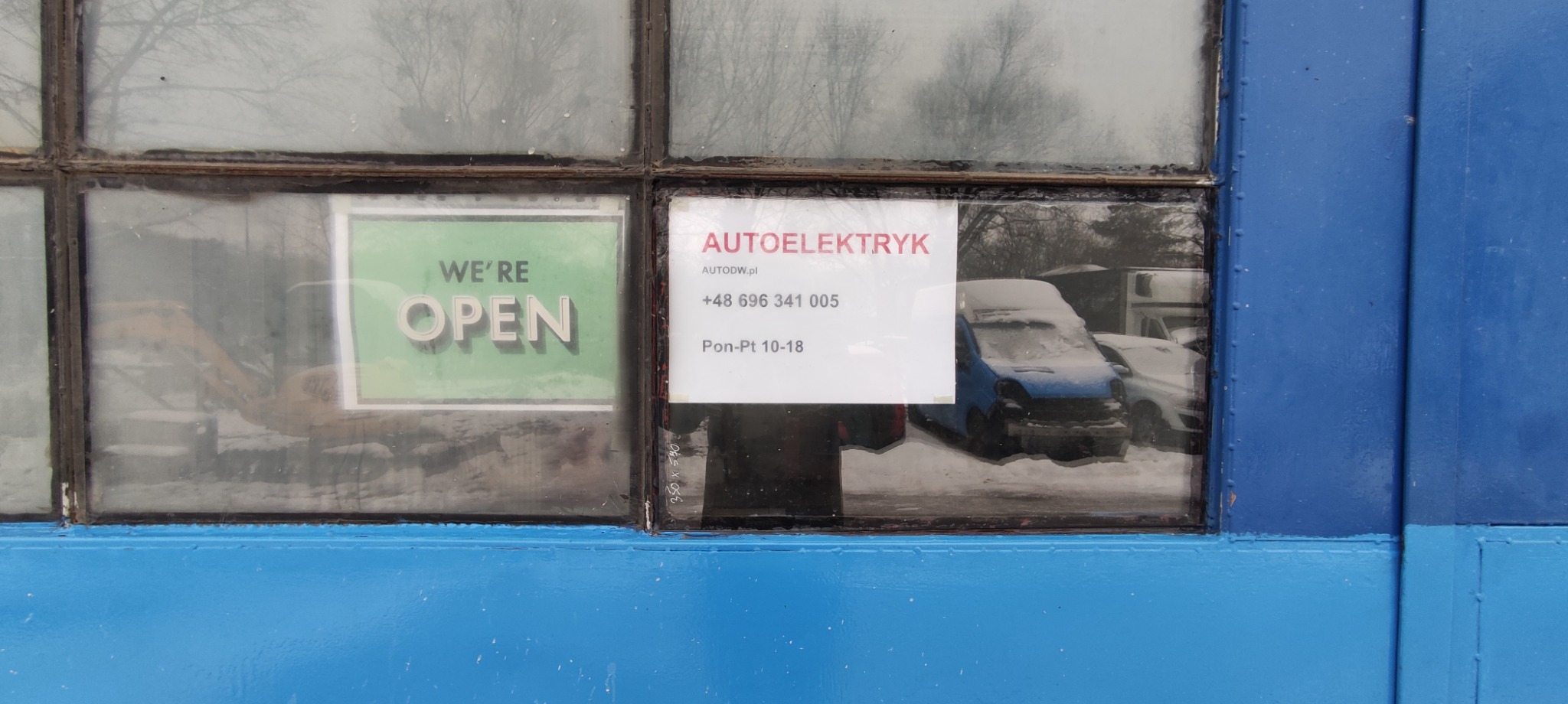 Szyba warsztatu z ogłoszeniem 'Jesteśmy otwarci' i kartką 'Autoelektryk', za szybą zaparkowane, zasypane śniegiem samochody.