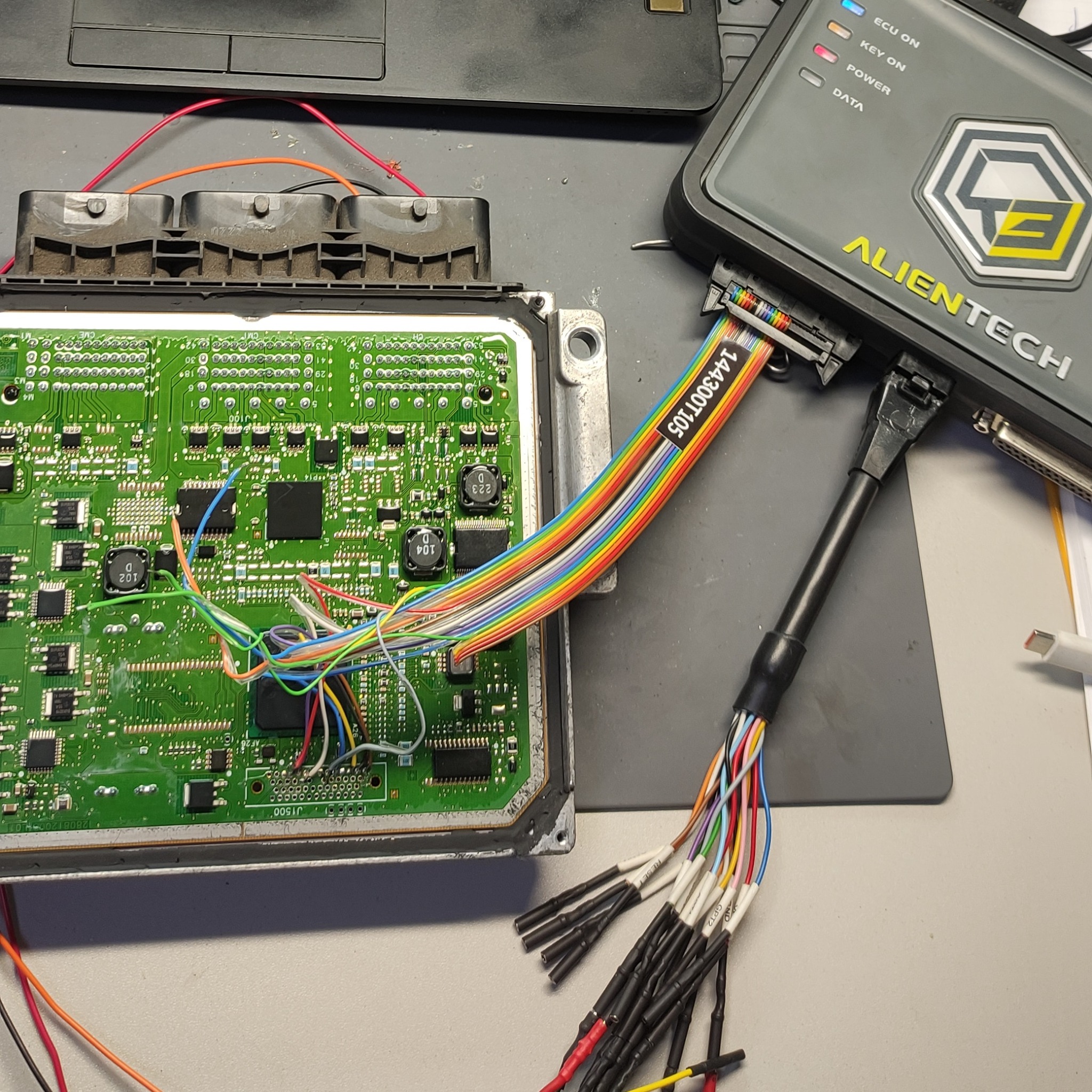 Otwarty moduł sterujący silnika (ECU) z podłączonymi kolorowymi przewodami do programatora Alientech, widoczne diody sygnalizacyjne urządzenia diagnostycznego oraz elementy elektroniczne na płytce...