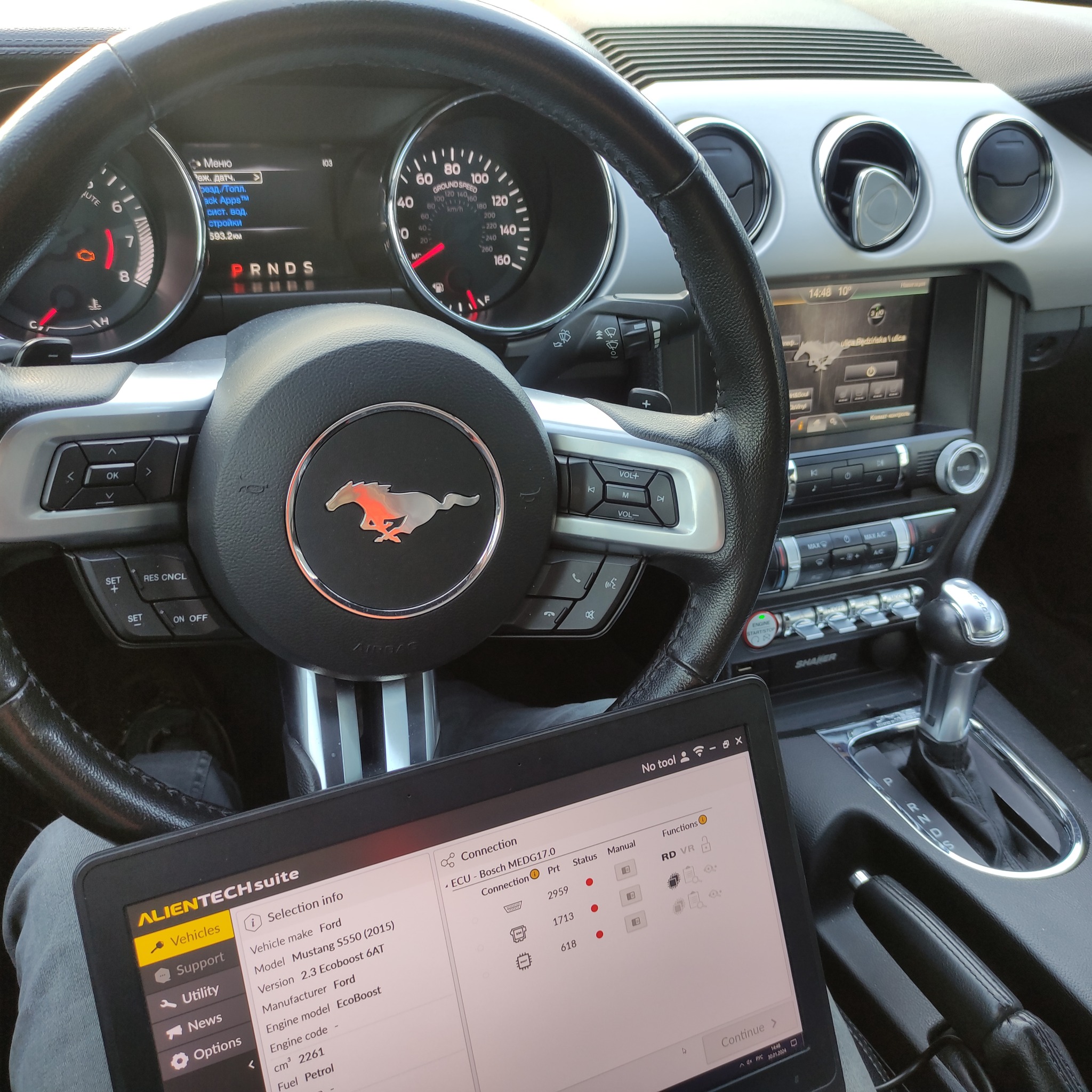 Diagnostyka komputerowa Ford Mustang S550 (2015) 2.3 EcoBoost. Tablet z uruchomionym oprogramowaniem Alientech Suite podłączony do ECU, widoczny kokpit i kierownica z logo Mustanga.