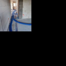 Kamil Wojtalik Klarvent - Niebieskie rury karbowane ułożone na podłodze w surowym wnętrzu podczas montażu instalacji, widoczne ściany z betonu komórkowego i oznaczenia instalacyjne.