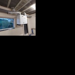 Kamil Wojtalik Klarvent - Biała jednostka centralna z podłączonymi przewodami wentylacyjnymi, zamontowana na ścianie w surowym wnętrzu, widoczne rury i okno z widokiem na zieleń.