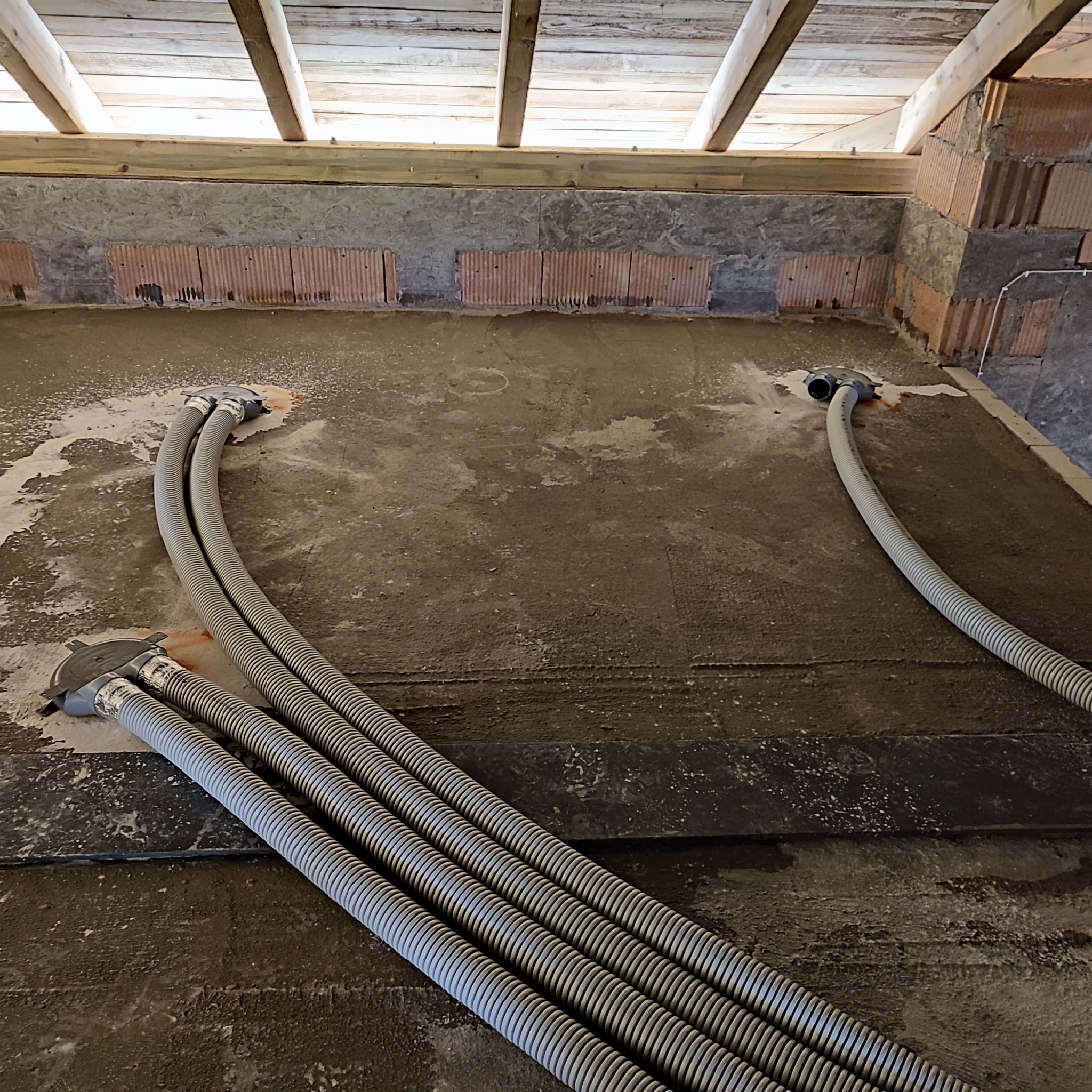 Szare, karbowane rury wentylacyjne ułożone na surowej, betonowej podłodze w trakcie montażu w budynku w stanie surowym, widoczne cegły i drewniana konstrukcja dachu.