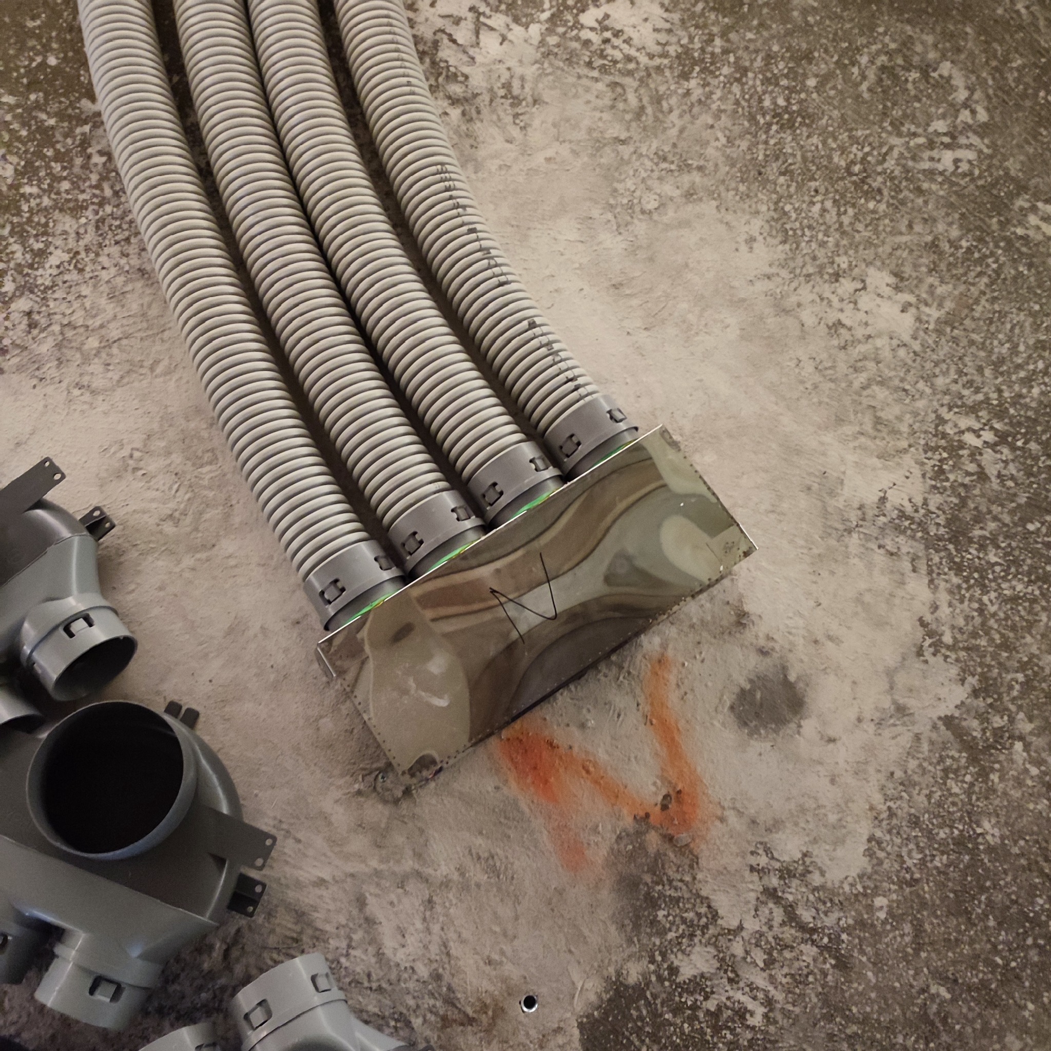 Cztery elastyczne rury wentylacyjne, połączone z metalową skrzynką rozdzielczą na surowym, szarym podłożu z pomarańczowymi oznaczeniami i elementami montażowymi.