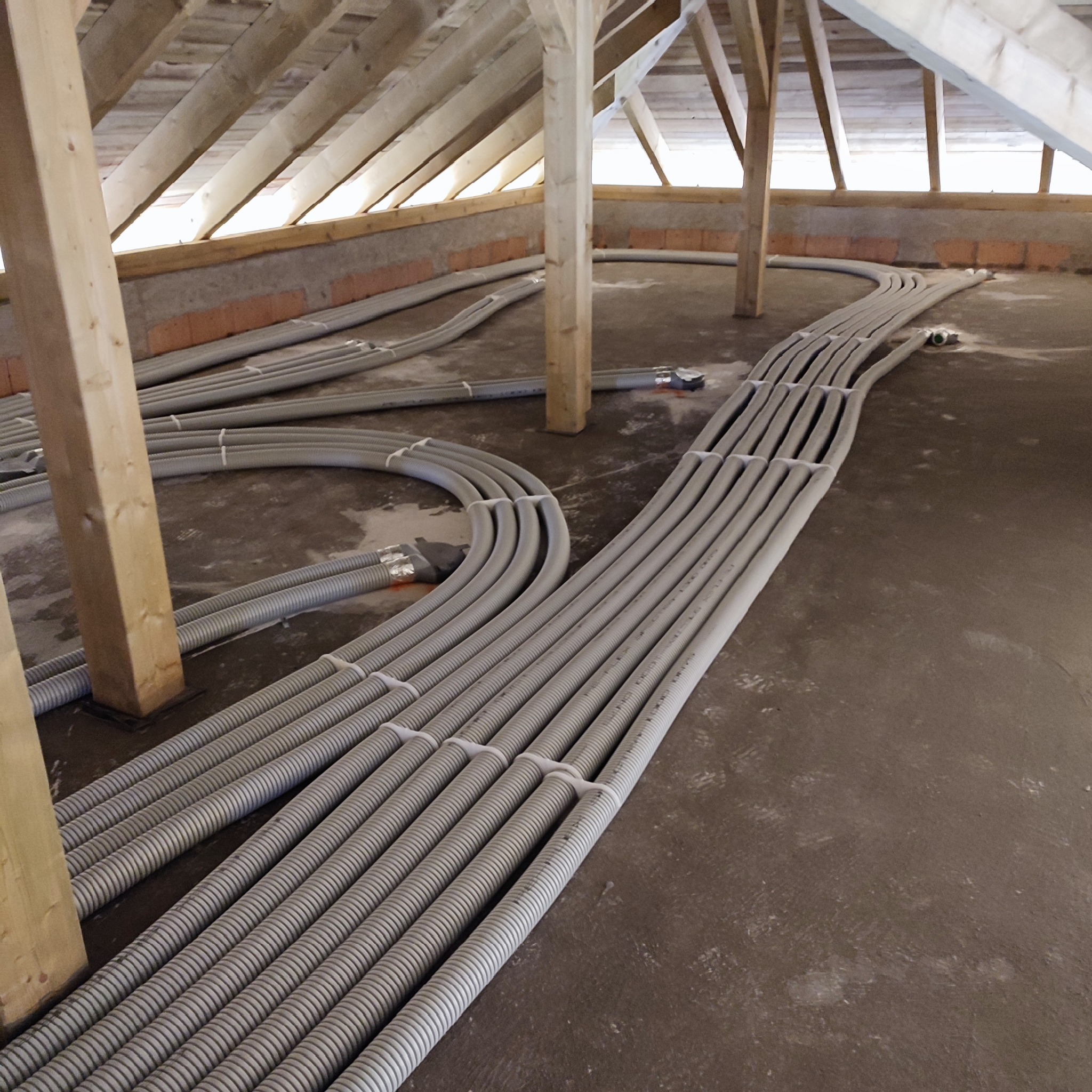 Szereg szarych, karbowanych rur wentylacyjnych ułożonych równolegle na betonowej podłodze poddasza, z widoczną drewnianą konstrukcją dachu i ceglanym murem w tle. Rury są połączone białymi opaskami...