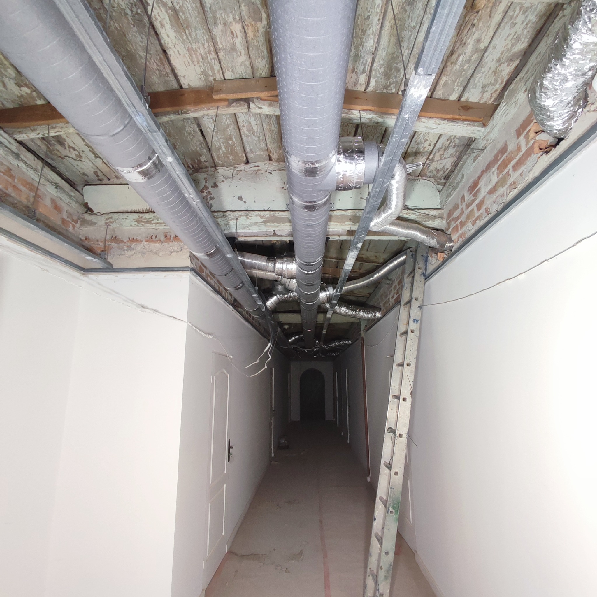Sufit korytarza z widoczną instalacją wentylacyjną z szarych, metalowych rur, częściowo izolowanych, zamontowanych do starego drewnianego stropu z resztkami farby, oraz drabina oparta o ścianę.