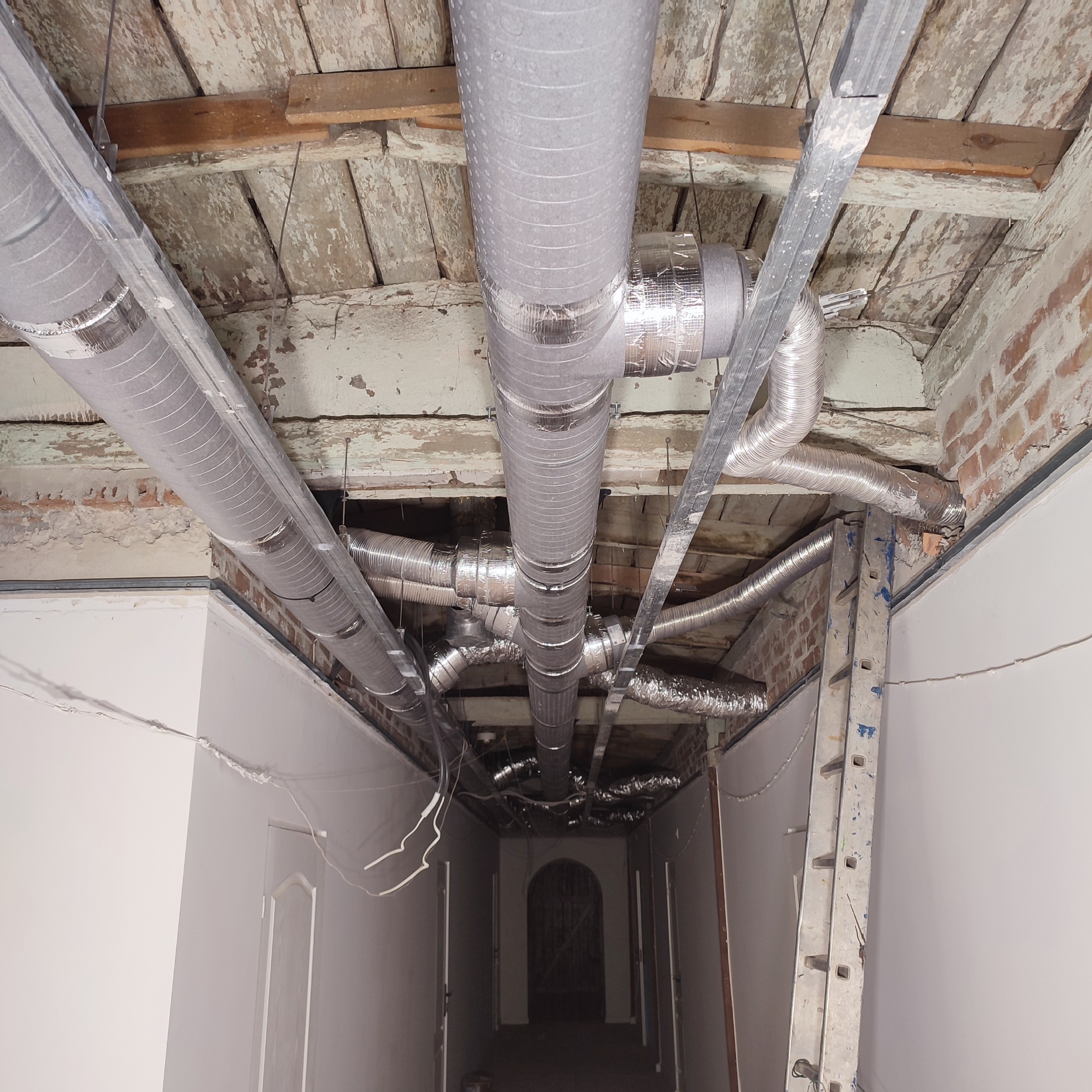 System kanałów wentylacyjnych z folii aluminiowej, zamontowany pod surowym sufitem z desek i odsłoniętej cegły w długim korytarzu podczas remontu.