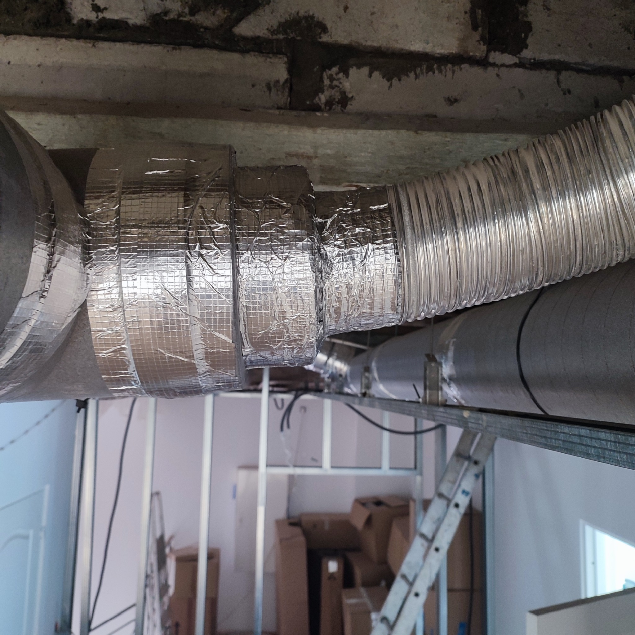 Połączenie dwóch przewodów wentylacyjnych, jeden elastyczny aluminiowy karbowany, drugi sztywny, owinięte srebrną taśmą izolacyjną, widoczne elementy konstrukcji podwieszanego sufitu i drabina w tle.