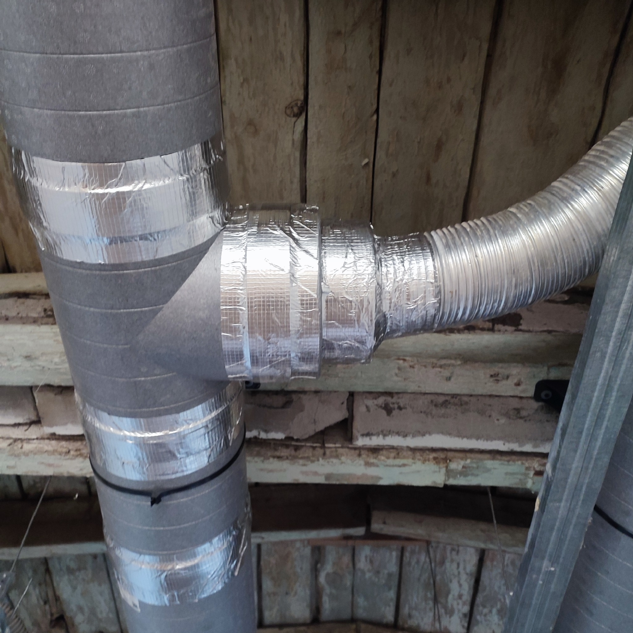Fragment instalacji wentylacyjnej z rurą spiro, zabezpieczoną taśmą aluminiową, widoczna na tle drewnianego stropu.