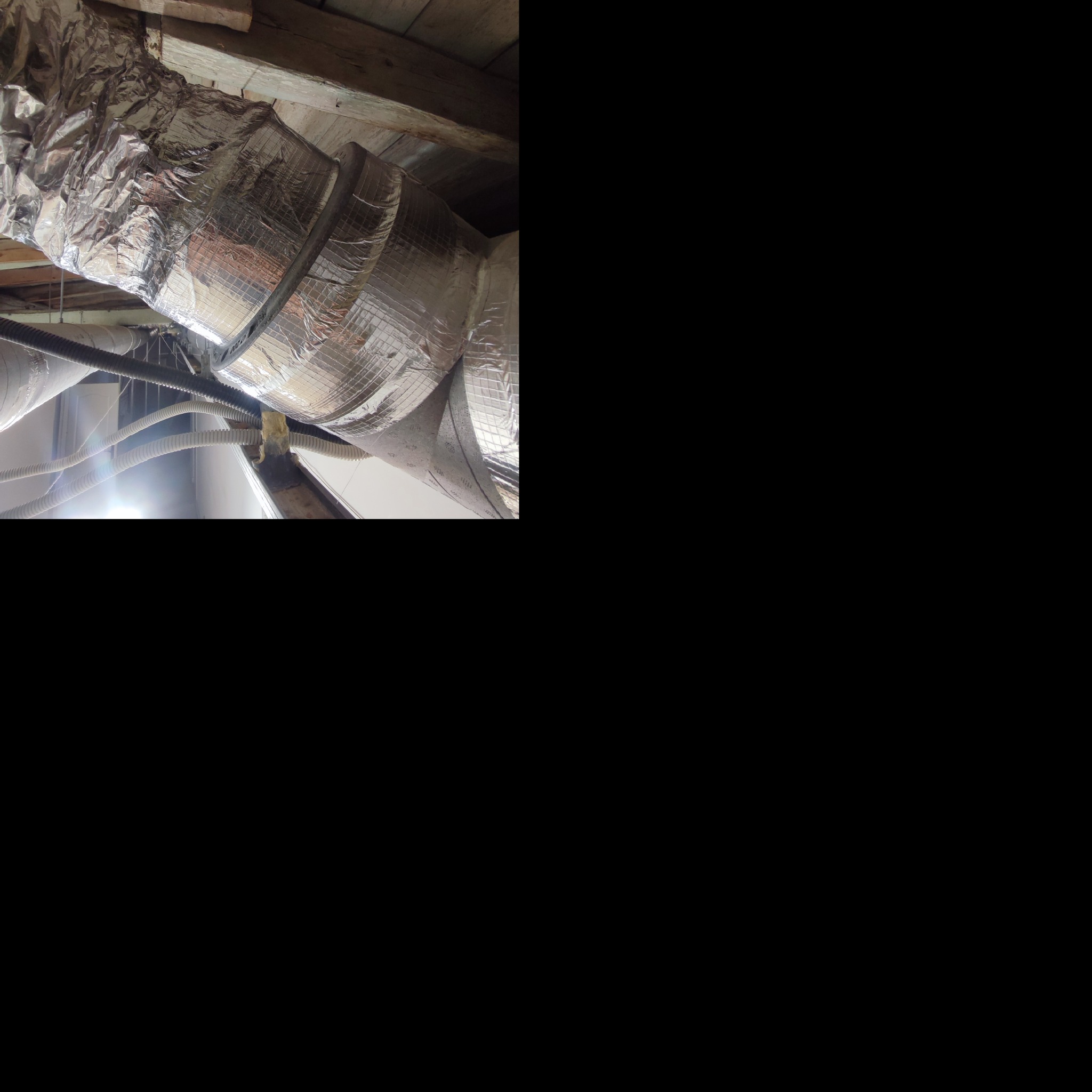 Srebrna, izolowana rura wentylacyjna przymocowana do drewnianego stropu, widoczne inne rury w tle.