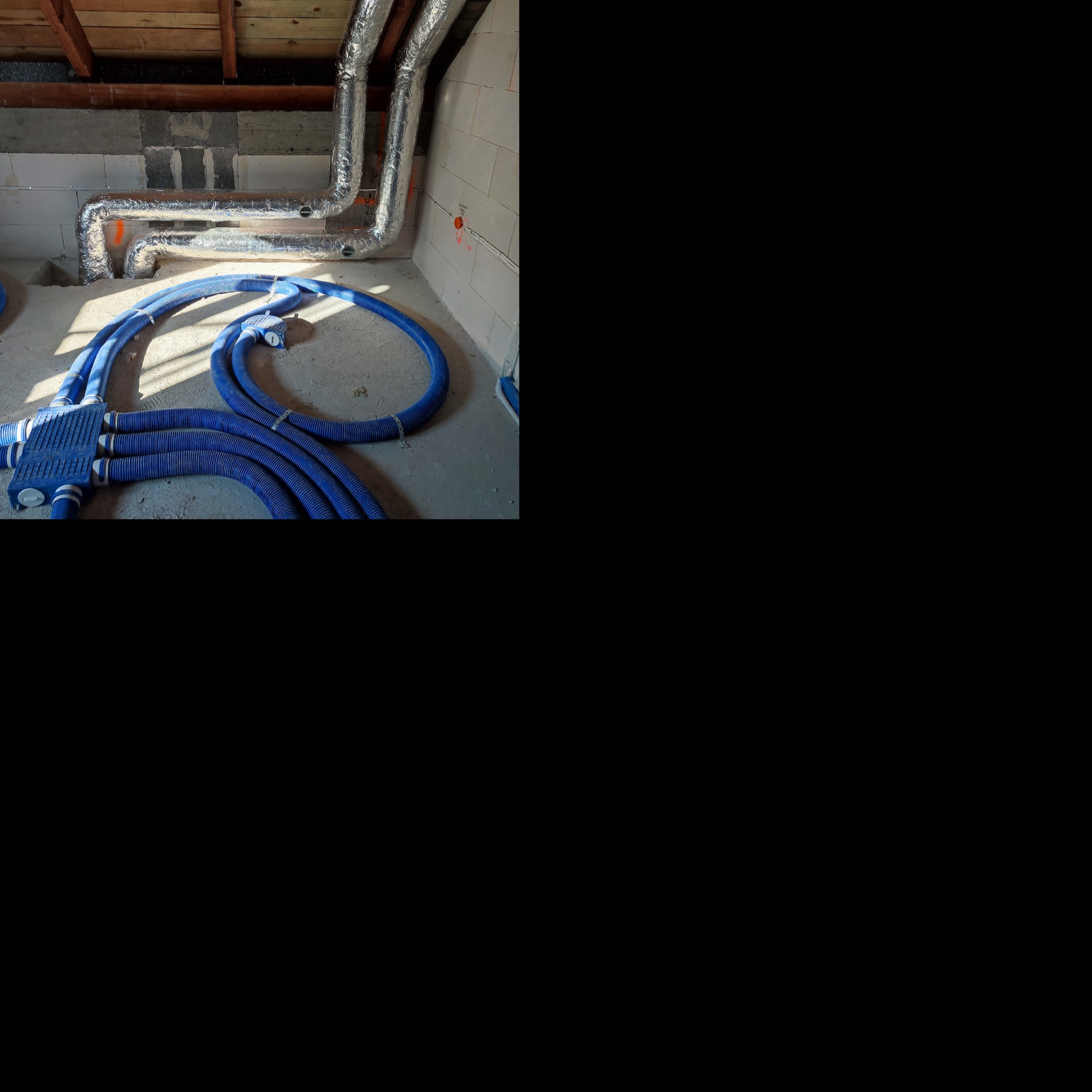 Rozdzielacz z podłączonymi niebieskimi rurami wentylacyjnymi na surowej podłodze, widoczne przewody w izolacji aluminiowej pod stropem.