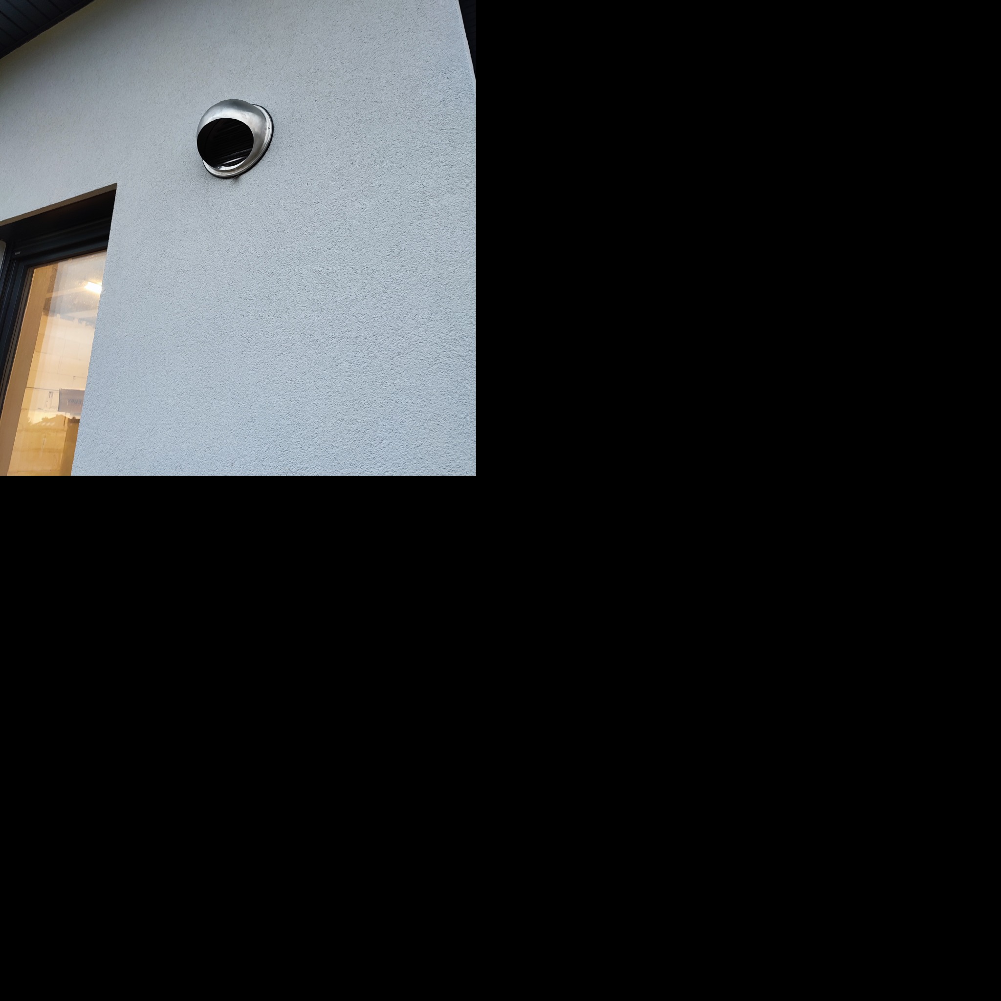 Metalowy anemostat na elewacji budynku z oknem, widoczne odbicie światła w metalu.