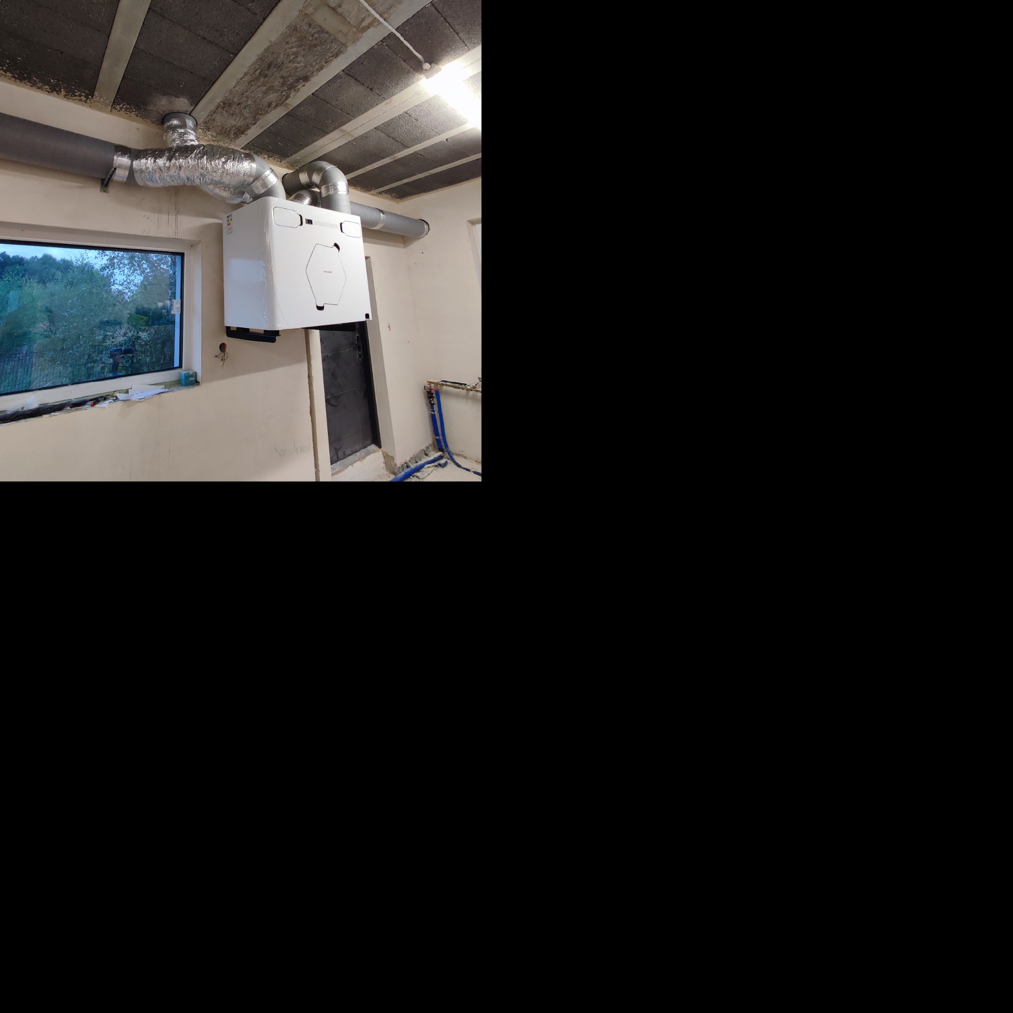 Biała jednostka centralna z podłączonymi przewodami wentylacyjnymi, zamontowana na ścianie w surowym wnętrzu, widoczne rury i okno z widokiem na zieleń.