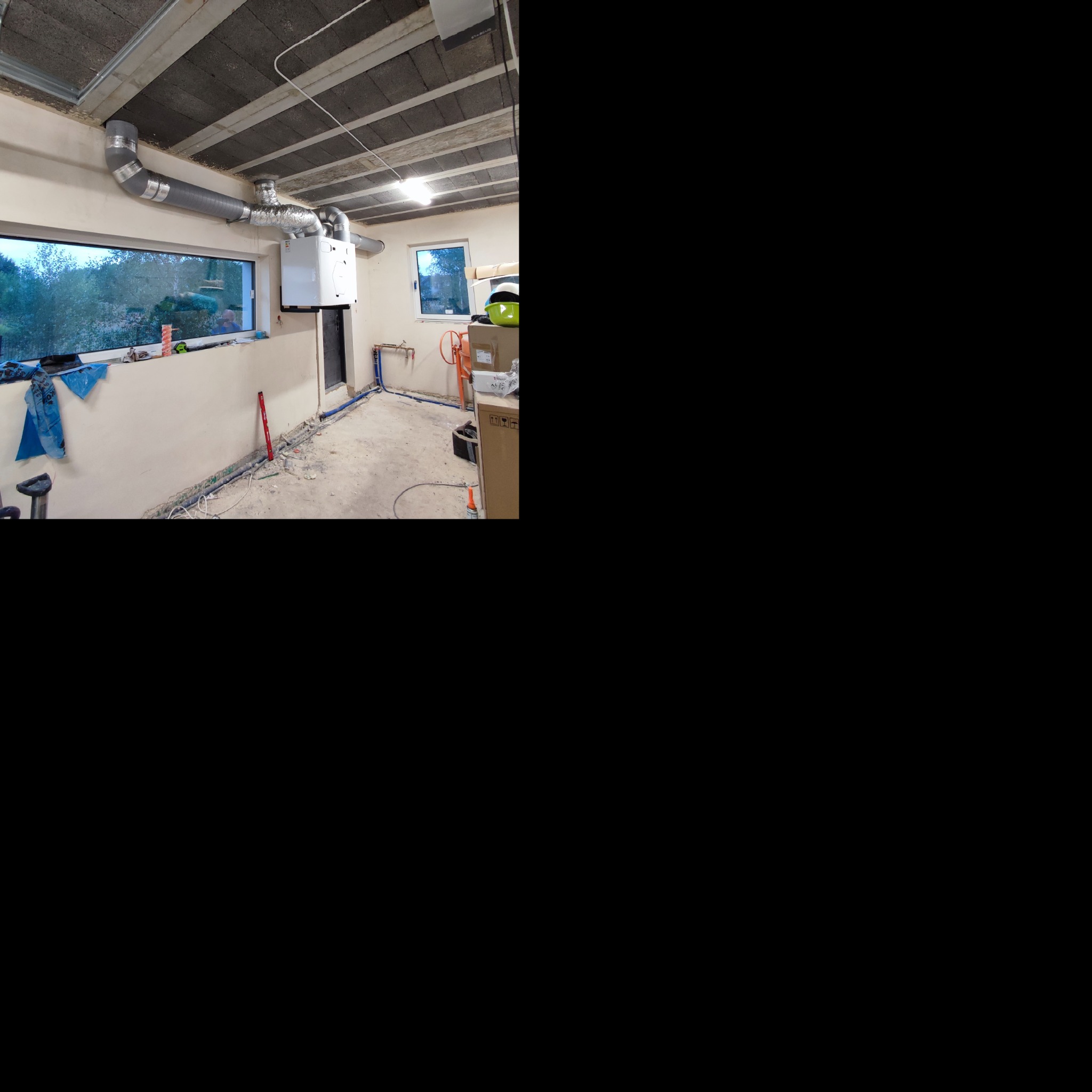 Szare rury wentylacyjne pod sufitem w surowym pomieszczeniu, biały piec wiszący na ścianie, widoczne instalacje wodne i narzędzia w tle.