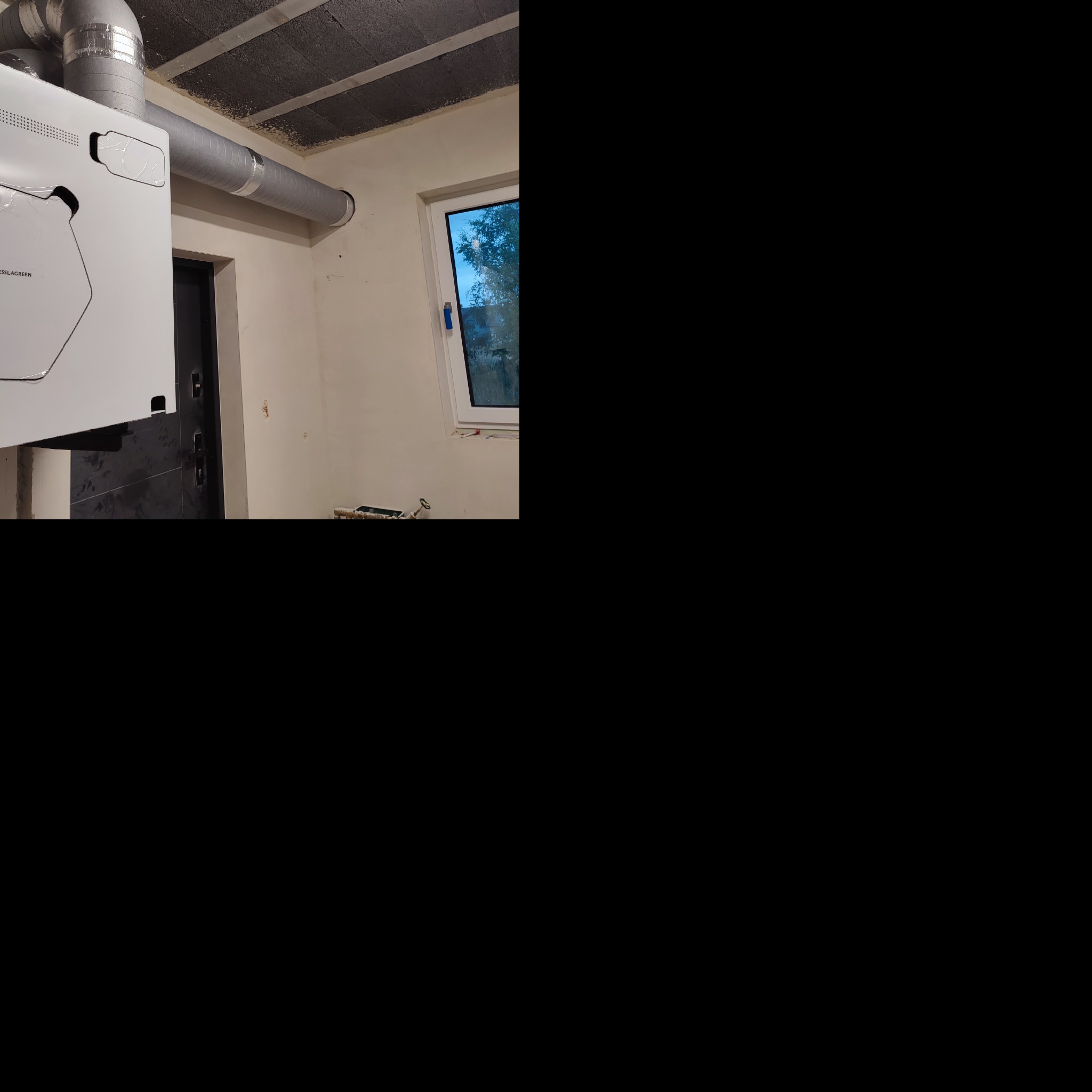 Biała jednostka centralna z podłączonymi srebrnymi rurami wentylacyjnymi w surowym pomieszczeniu z oknem i drzwiami.