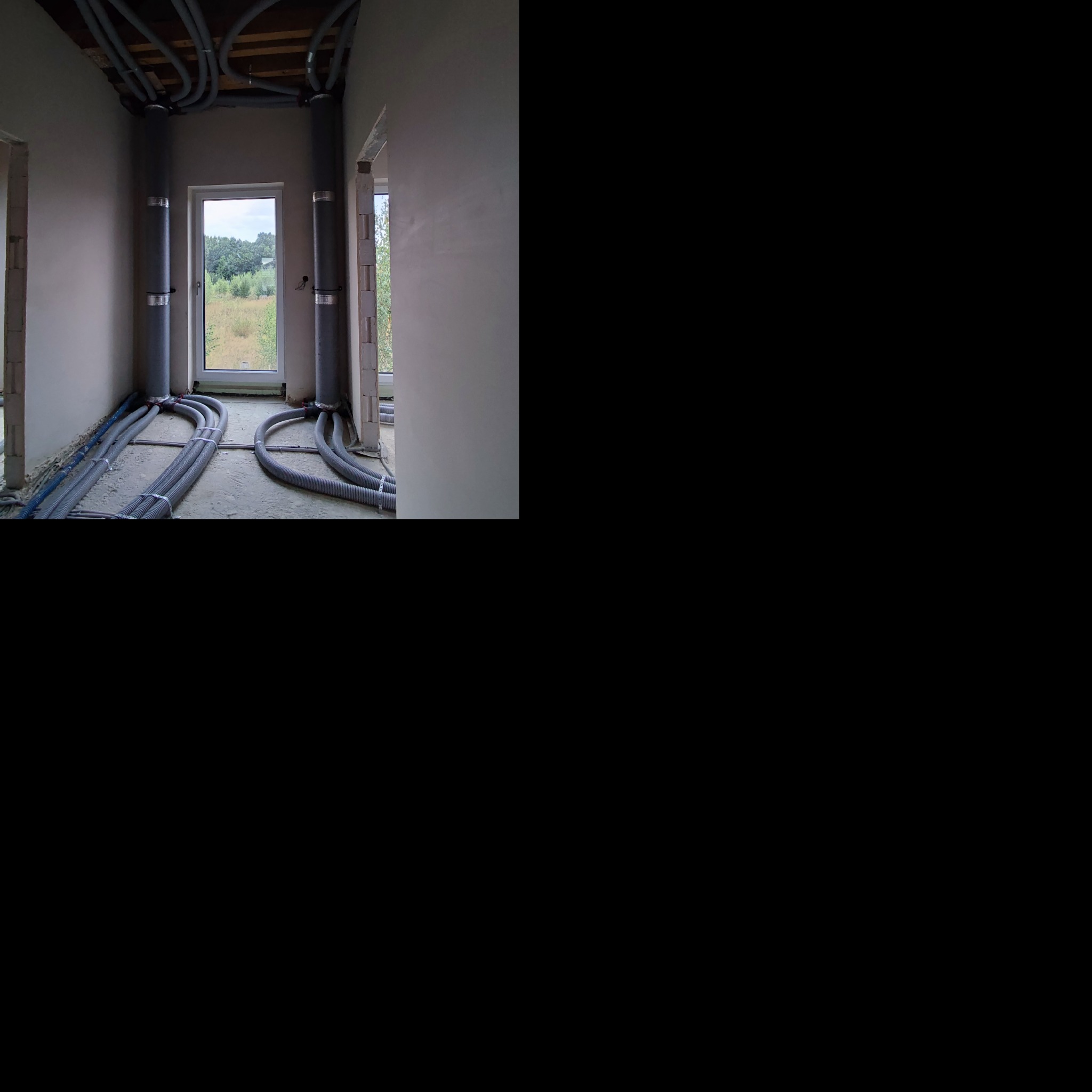 Szare rury wentylacyjne i instalacyjne prowadzone po podłodze i pod sufitem w surowym wnętrzu budynku w trakcie budowy, widoczne okno z widokiem na zieleń.