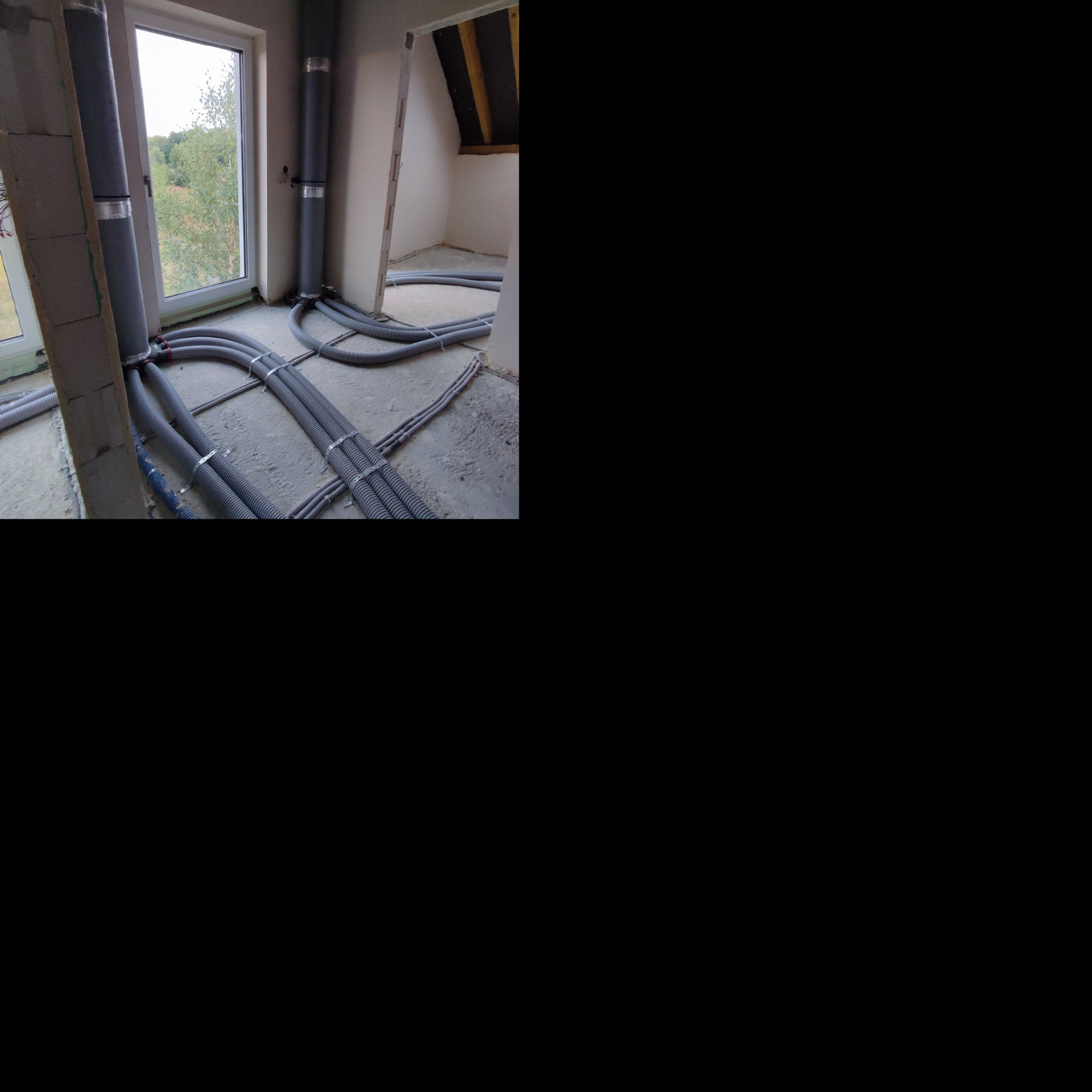 Szare, karbowane rury wentylacyjne pionowe i poziome, zabezpieczone metalowymi opaskami, rozprowadzone w surowym pomieszczeniu z oknem, podczas instalacji.