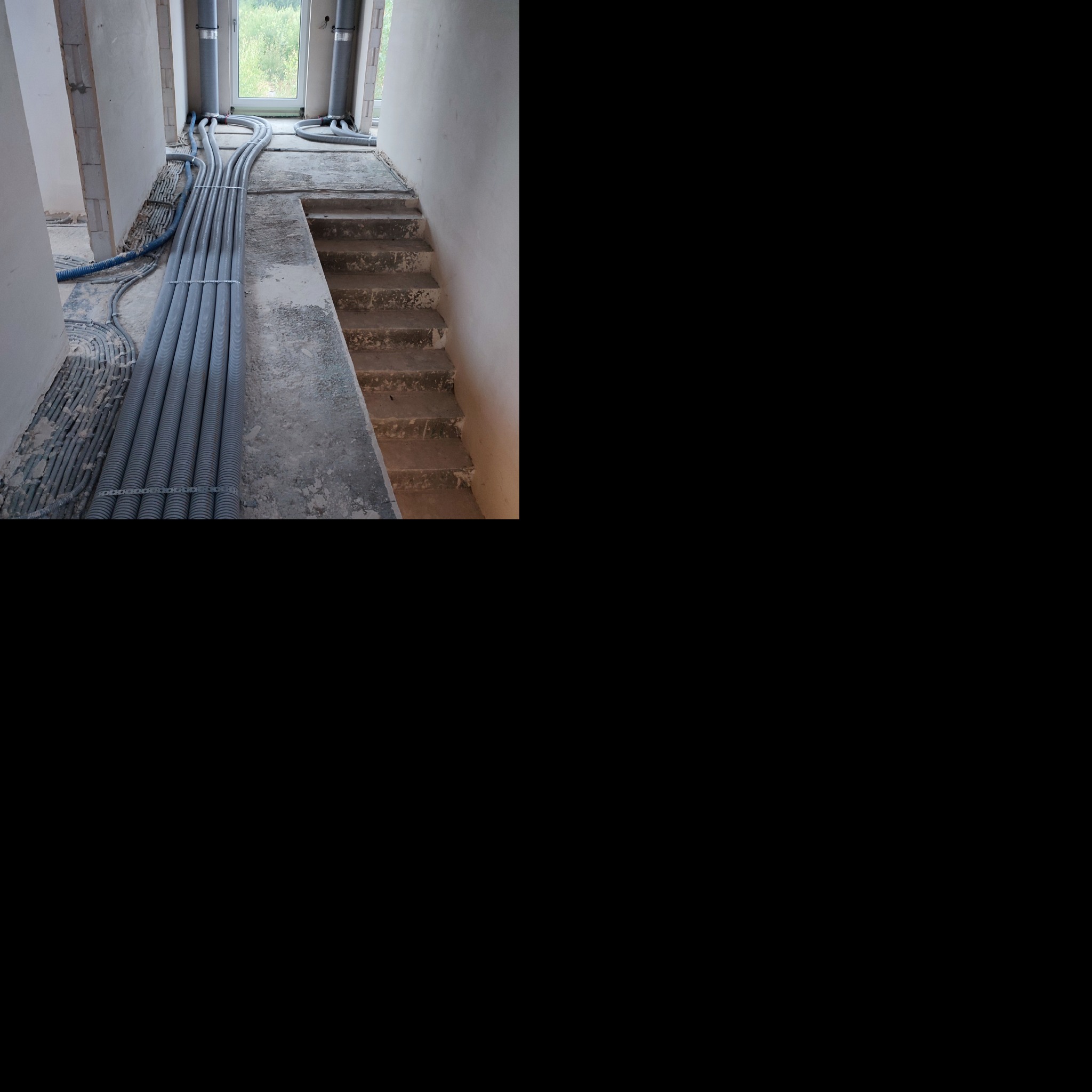 Szereg szarych, karbowanych rur wentylacyjnych ułożonych równolegle na betonowej podłodze w surowym wnętrzu budynku w trakcie budowy, z widocznymi schodami i oknami w tle.