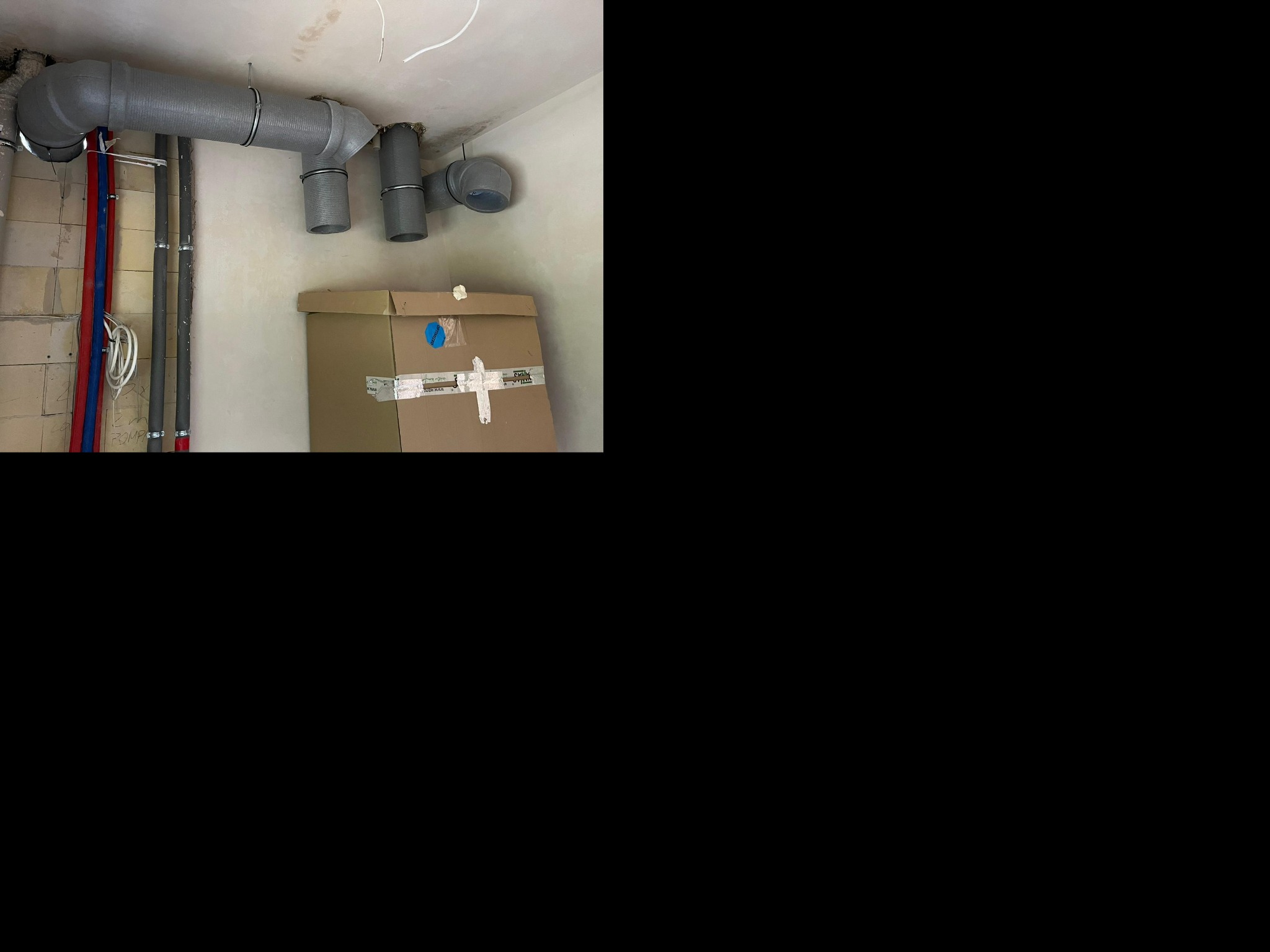 Szare rury wentylacyjne pod sufitem w surowym wnętrzu, widoczne pionowe rury instalacyjne w kolorze czerwonym i niebieskim, oraz kartonowe pudło pod instalacją.